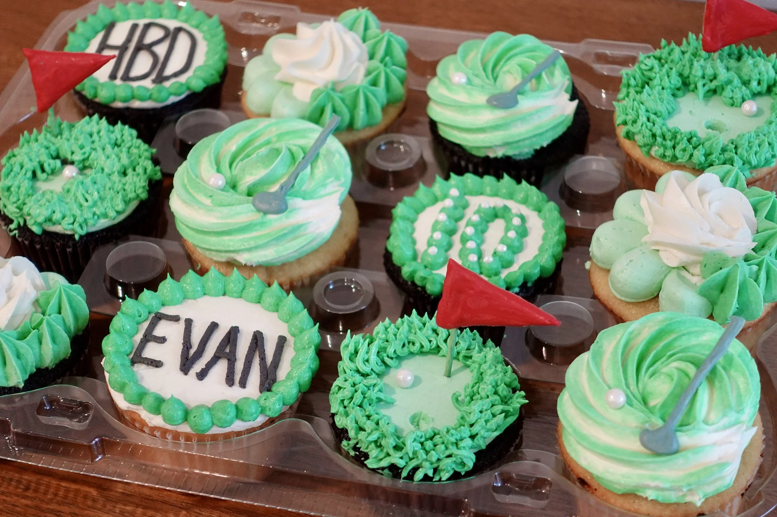 A collection of cupcakes decorated with green, white, and black icing, some with small flags, pearls, and a sign reading 'HBD'.