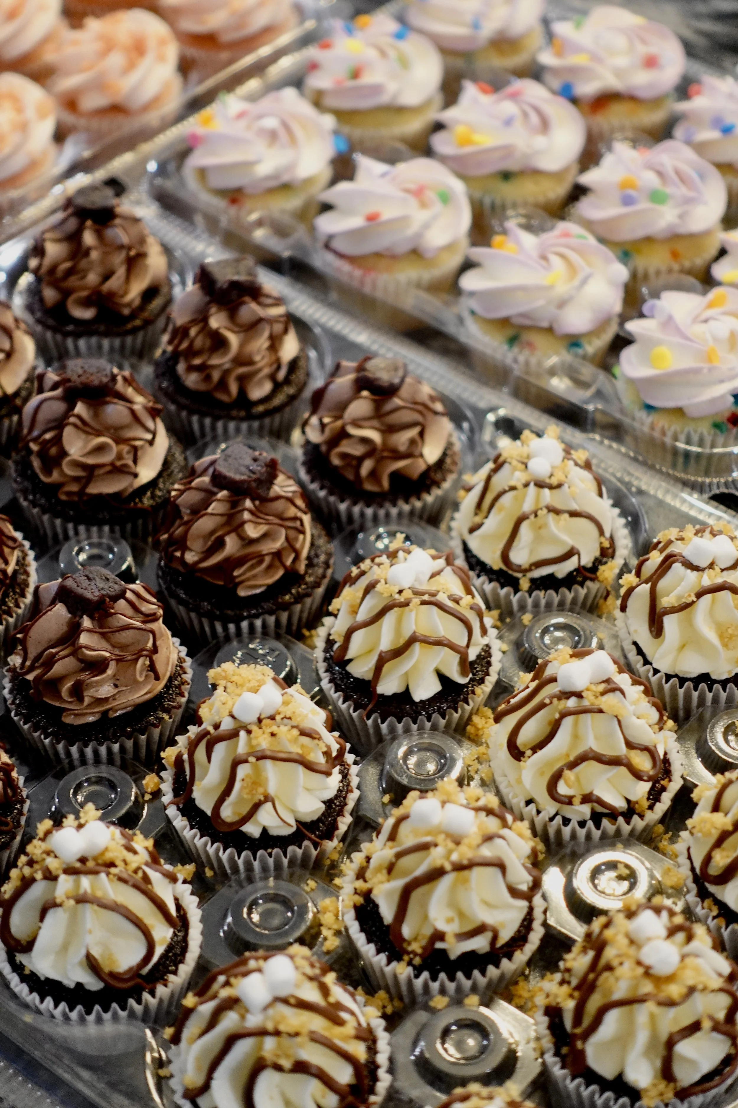 A tray of assorted cupcakes with various frosting decorations and toppings including sprinkles, chocolate drizzle, and crumbled cookies.