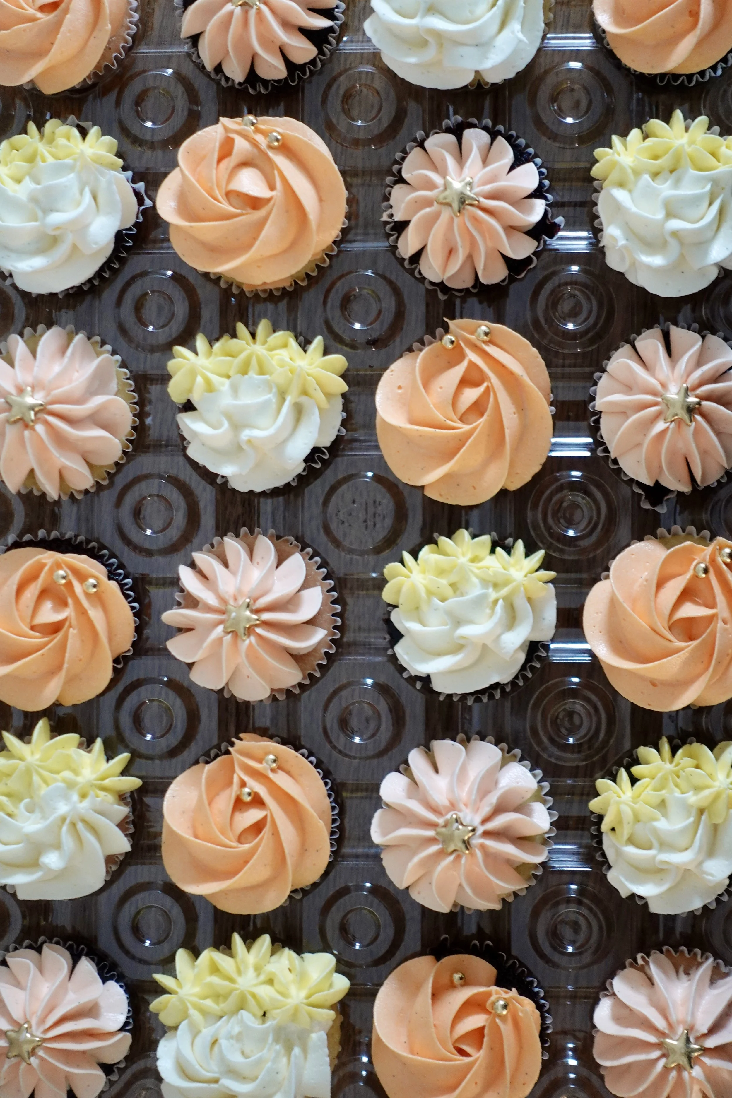 Assorted cupcakes decorated with pastel-colored frosting in peach, cream, and yellow, some with metallic star or ball toppers. The cupcakes are arranged in a grid on a clear plastic tray.