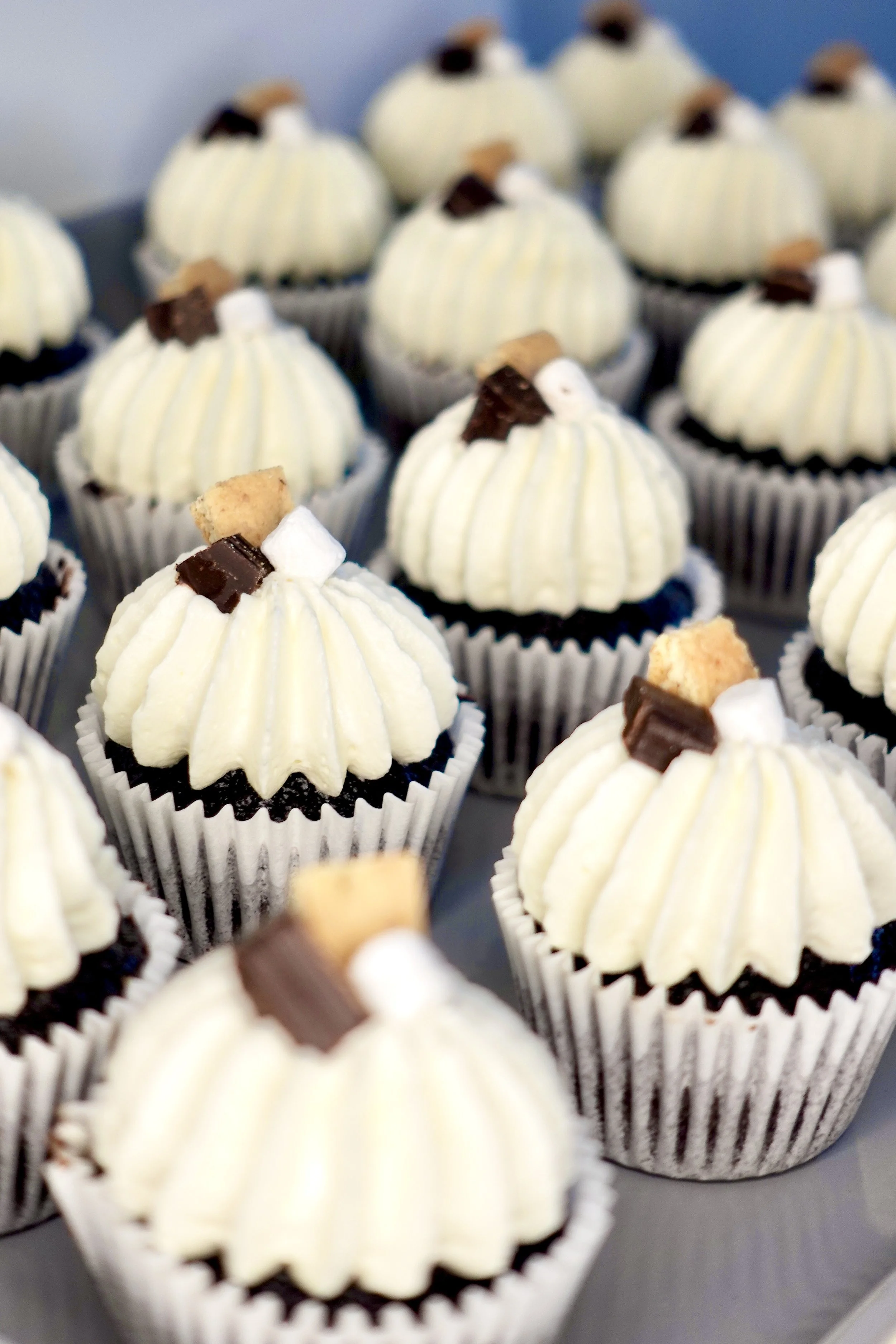 A group of cupcakes with white frosting decorated with small pieces of chocolate, cookie crumbs, and mini marshmallows, in black and white striped cupcake liners.