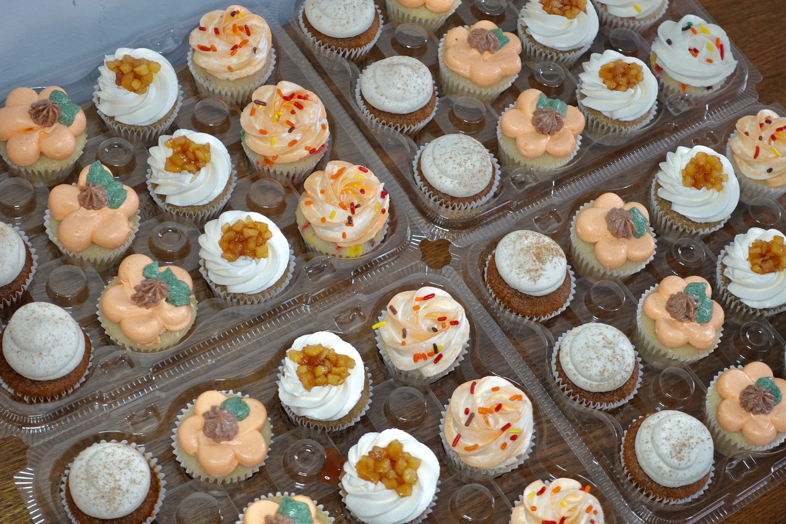 Assorted decorated cupcakes in a clear plastic container, topped with icing, sprinkles, and fruit compote.