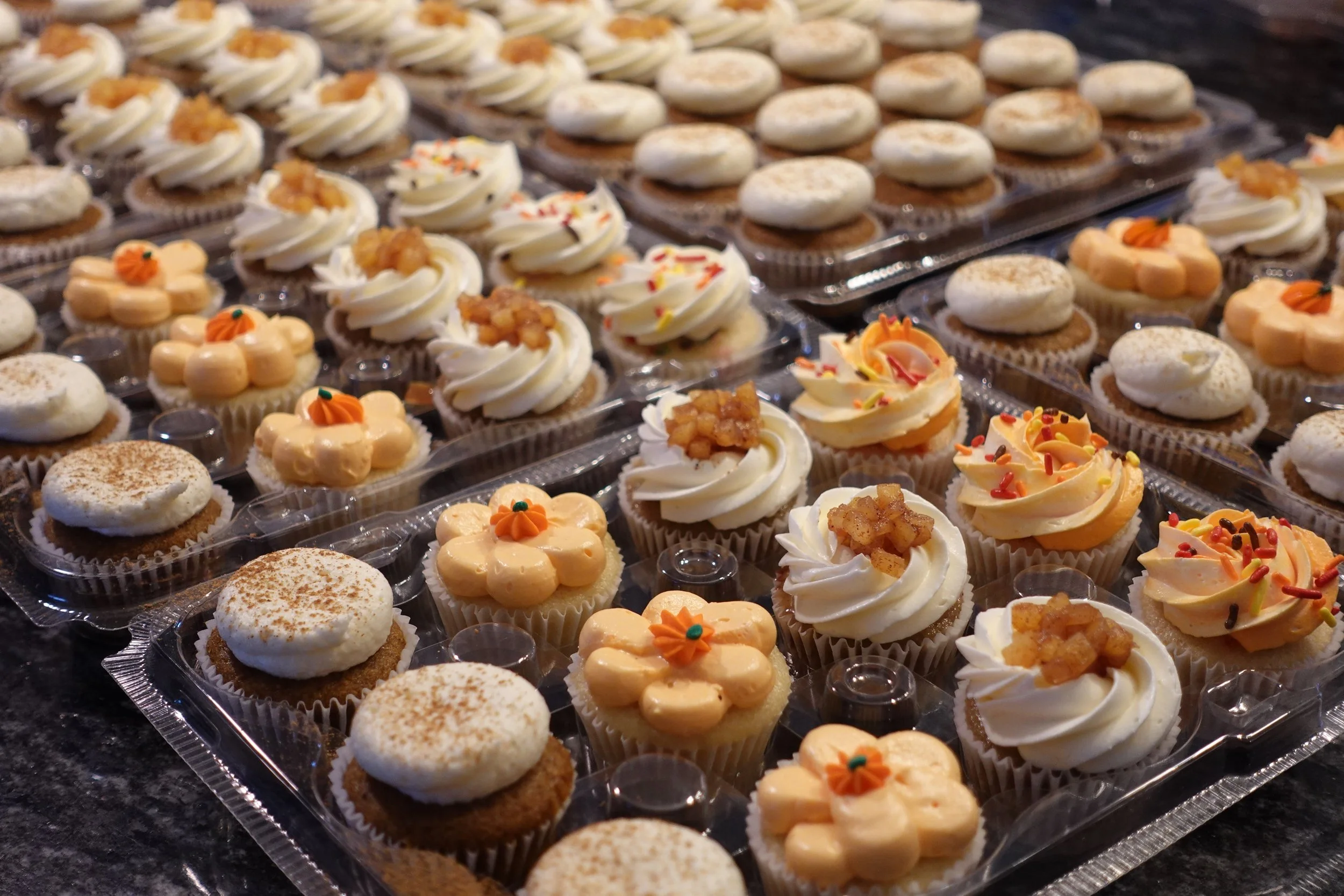 Assorted cupcakes on display, decorated with swirls of frosting, sprinkles, and small decorative flowers, arranged in clear plastic trays on a dark surface.