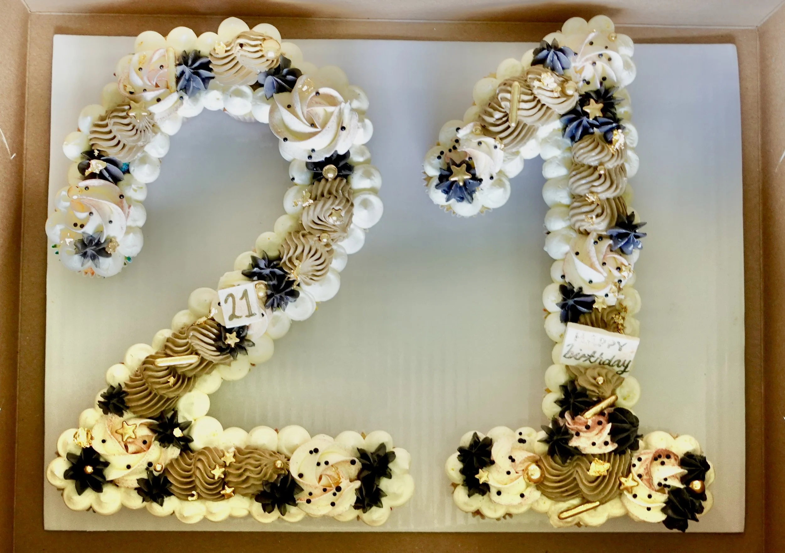 A birthday cake shaped like the number 21, decorated with white, beige, and black frosting, star-shaped and pearl decorations, and a small sign with 'happy birthday' written on it.