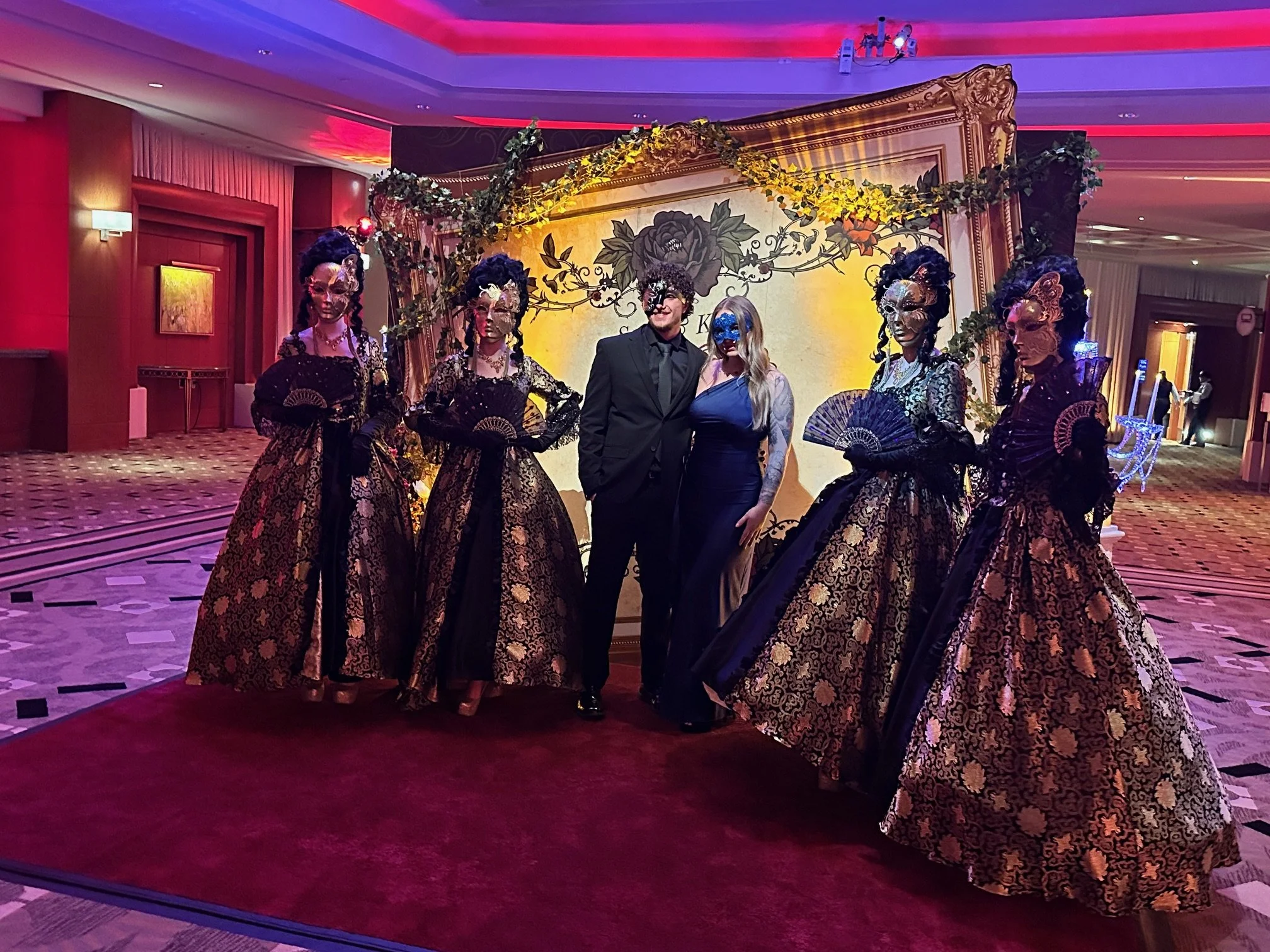 People dressed in elaborate costumes with masks, standing in front of a decorative backdrop at an event or celebration in a hotel or banquet hall.