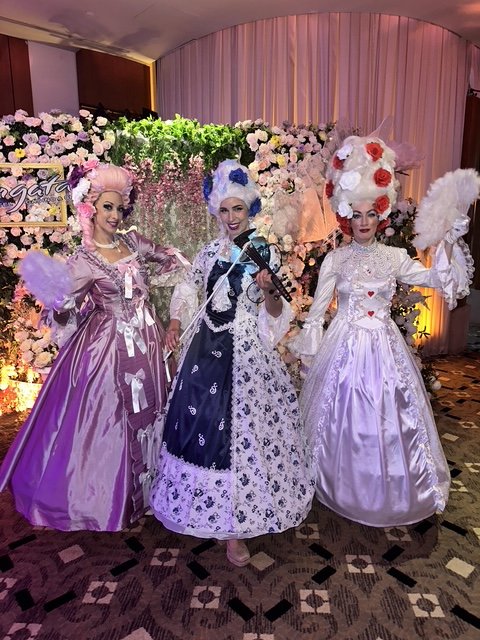 Three women dressed in elaborate 18th-century style, Bridgerton inspired gowns and wigs, standing in front of a floral backdrop at a New Year's Eve  event in a casino.