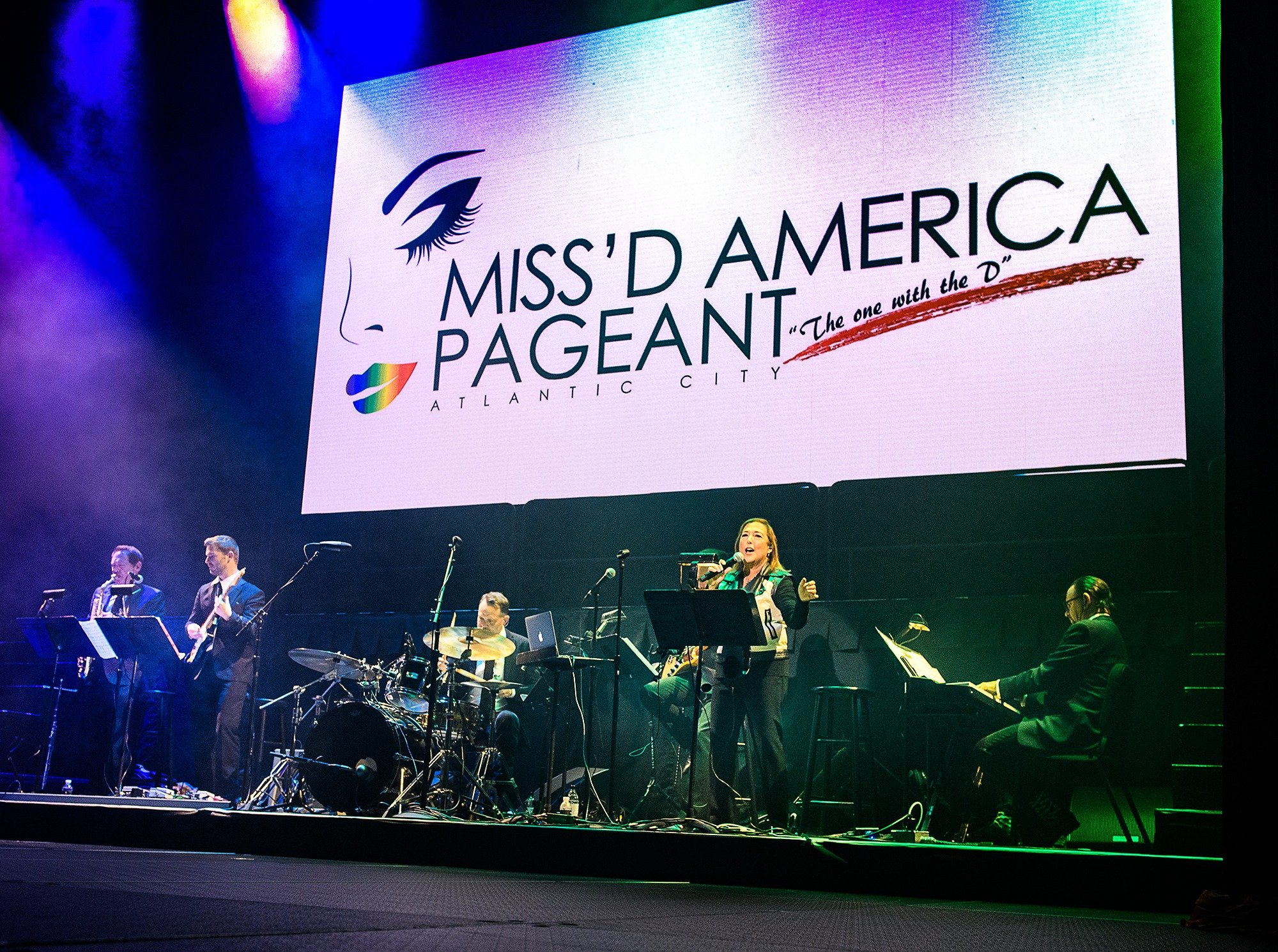 A concert stage with a large screen displaying the Miss'D America Pageant logo, featuring a stylized eye and a rainbow-colored heart, with the Melanie Rice Orchestra performing live music in front of the screen.