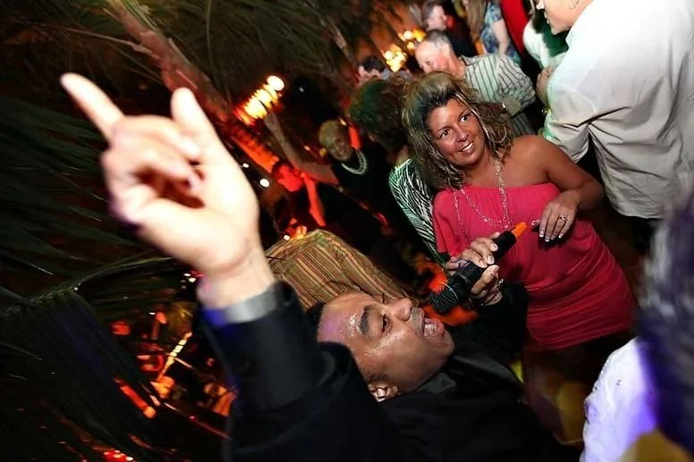 People dancing and socializing at an outdoor party or event during nighttime. One person in the foreground is dancing with their arm raised, while others are mingling and enjoying the lively atmosphere.