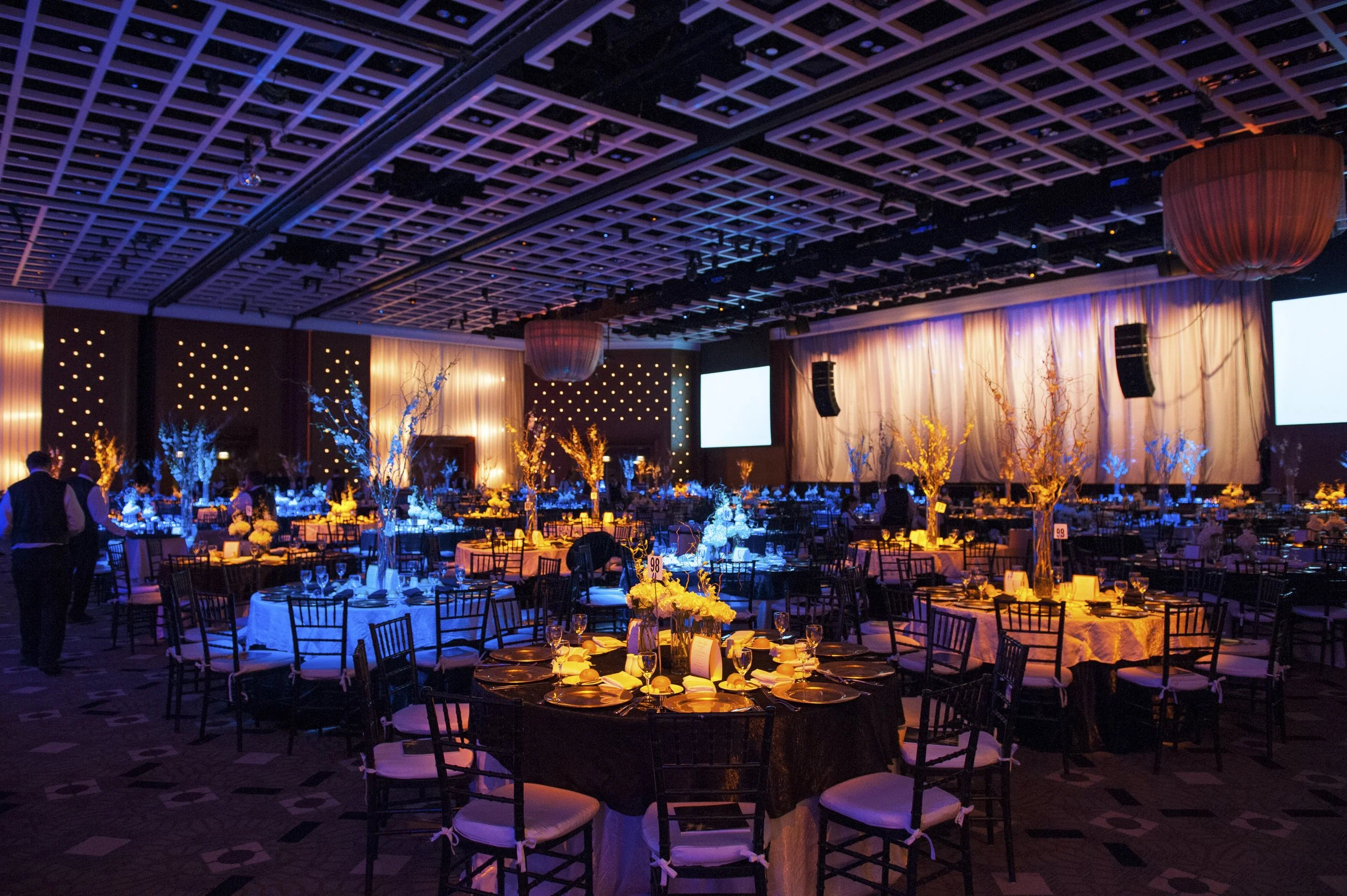 A large banquet hall decorated for an event with round tables, floral centerpieces, and chairs. The hall features high ceilings with a grid pattern and is illuminated with ambient and blue lighting. There are large screens and hanging lanterns.