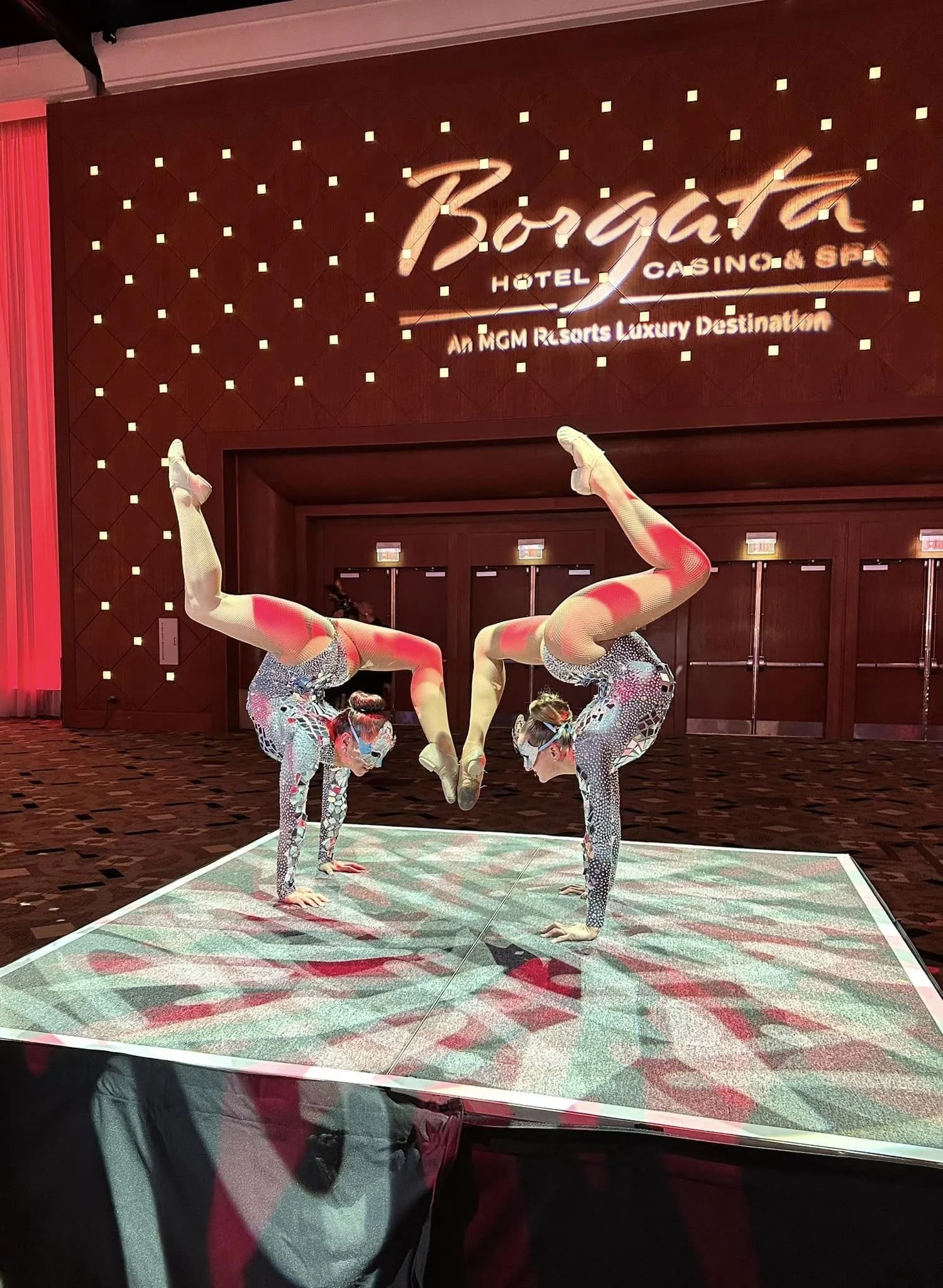 Two female contortionists performing a routine in a hotel ballroom event center room, with a large "Borgata" sign on the wall behind them.