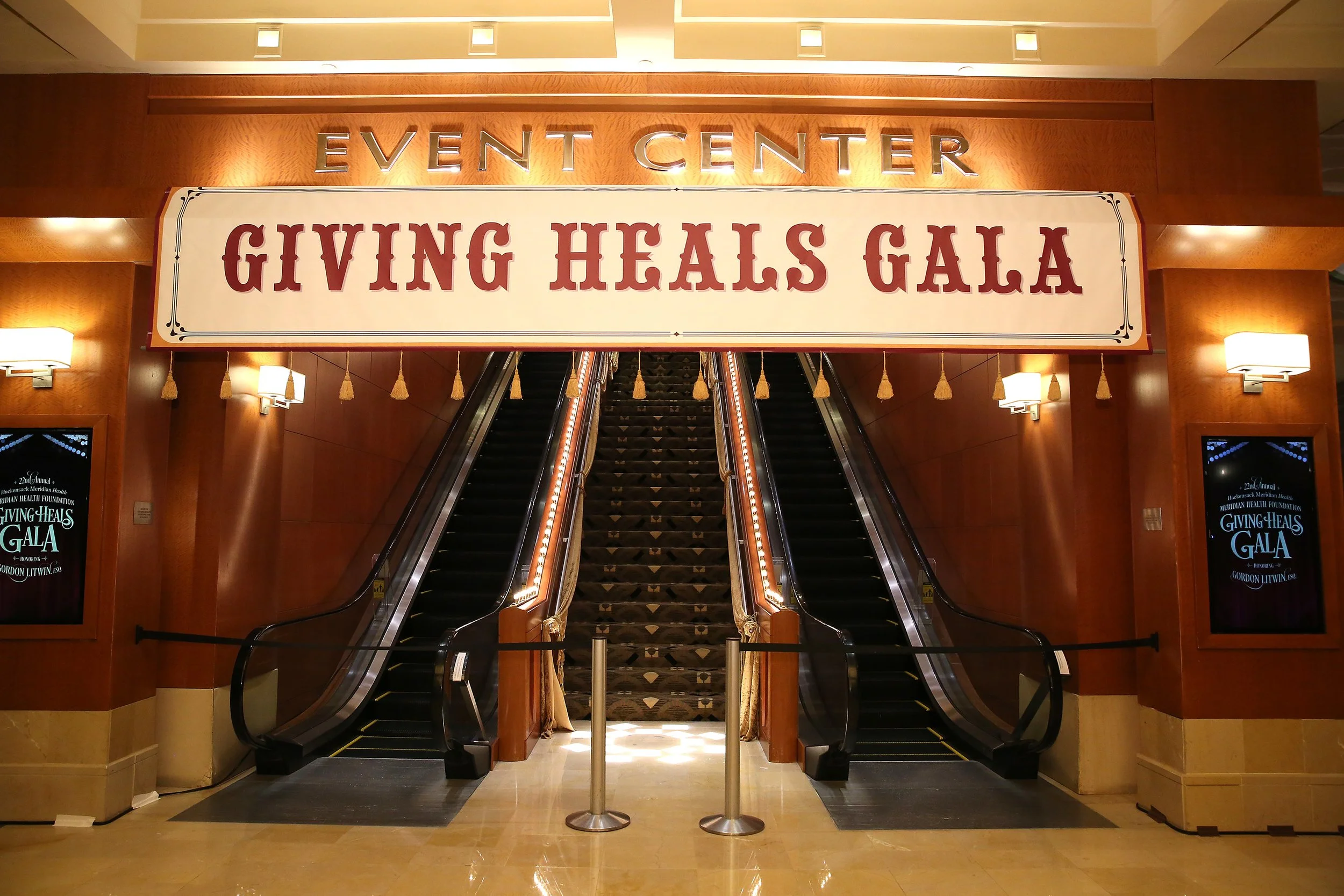Entrance to an event center for a Giving Heals Gala, featuring two escalators in a wooden-finished lobby area with decorative wall lamps and a large sign hanging above the escalators.