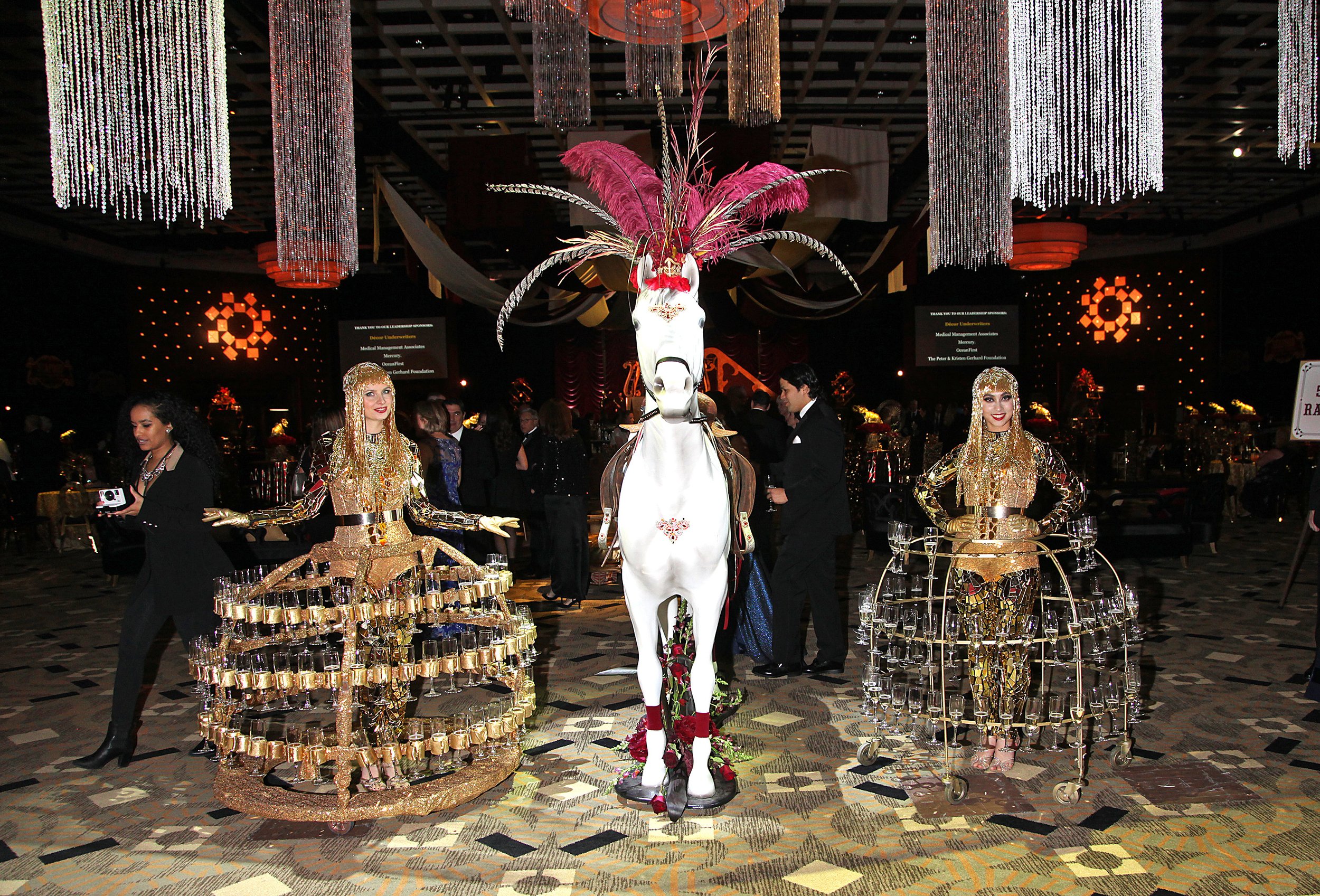 Guests at a Gala event with a white horse decor piece with a pink feather headdress at the center. The room is decorated with hanging crystal chandeliers and orange lighting, with two gold-colored champagne divas serving cocktails.