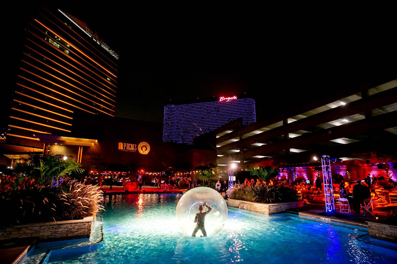 Night scene at a casino hotel with a lit pool, a contortionist inside a floating sphere on the pool, surrounded by illuminated buildings and outdoor seating areas.