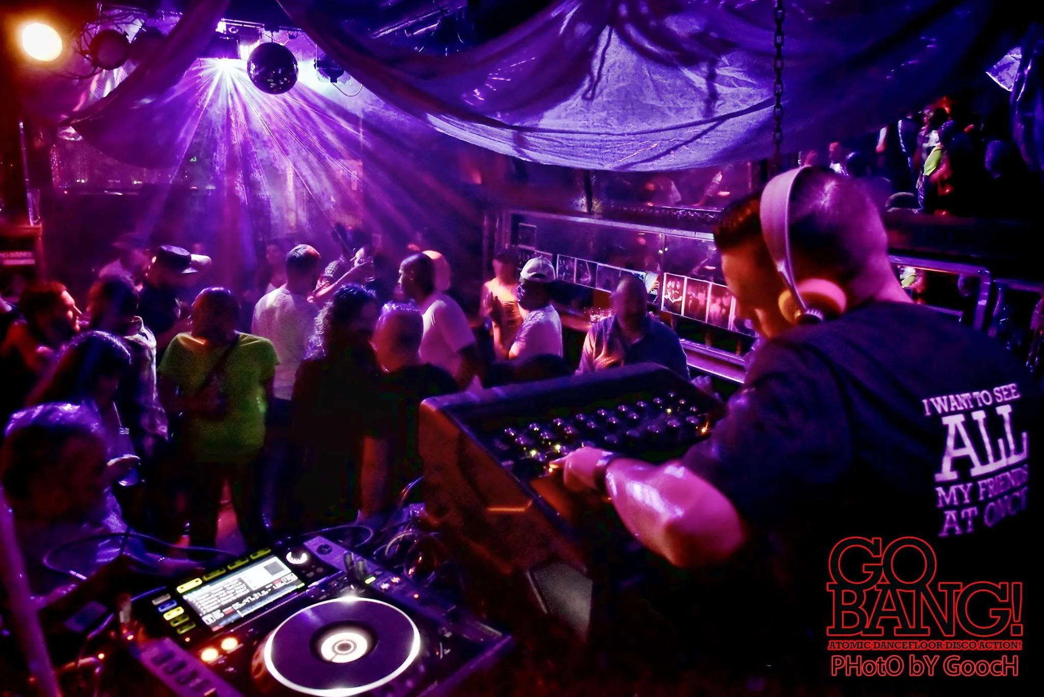 People dancing and partying in a nightclub to a DJ with purple and blue lighting, DJ equipment in the foreground, and a disco ball hanging from the ceiling.