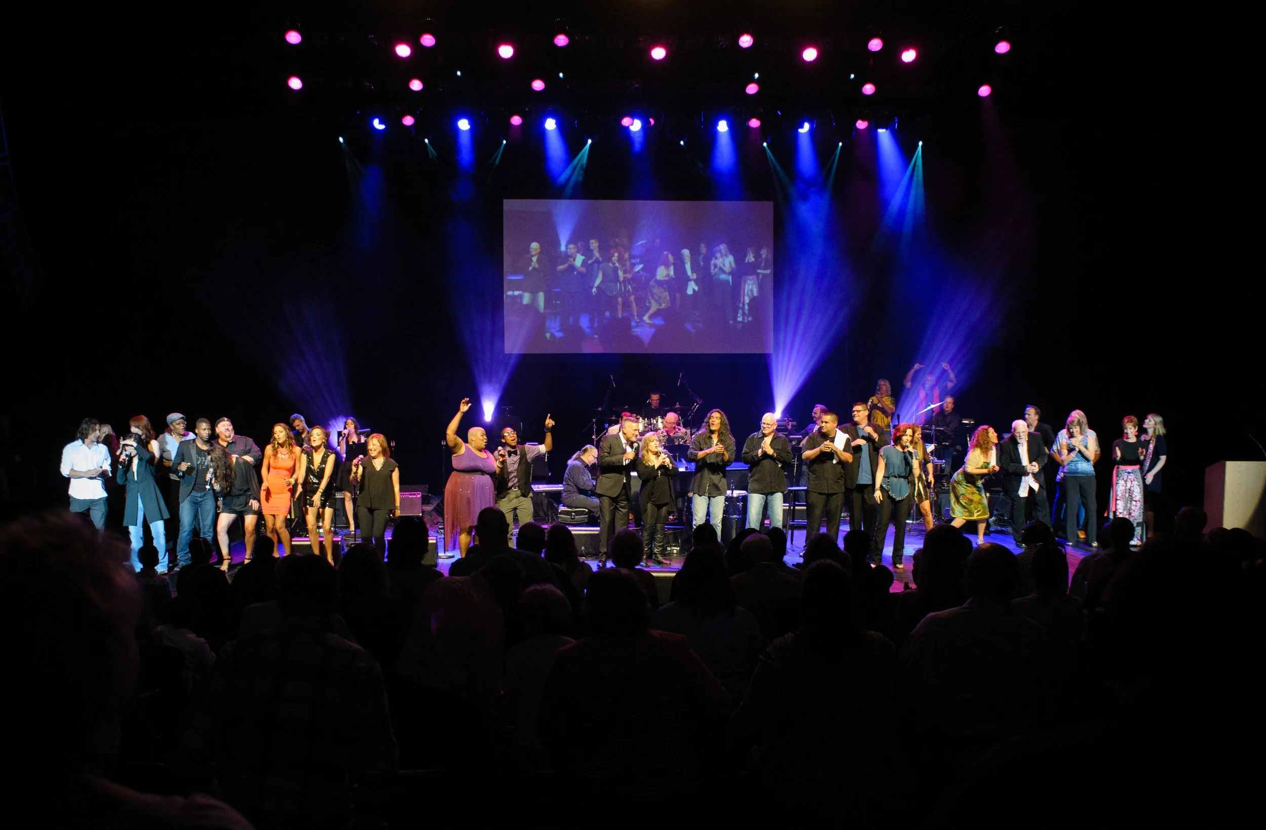 A large group of people on a concert stage performing or speaking at an event, with colorful stage lighting and a projection screen displaying a reflection of the group above them.