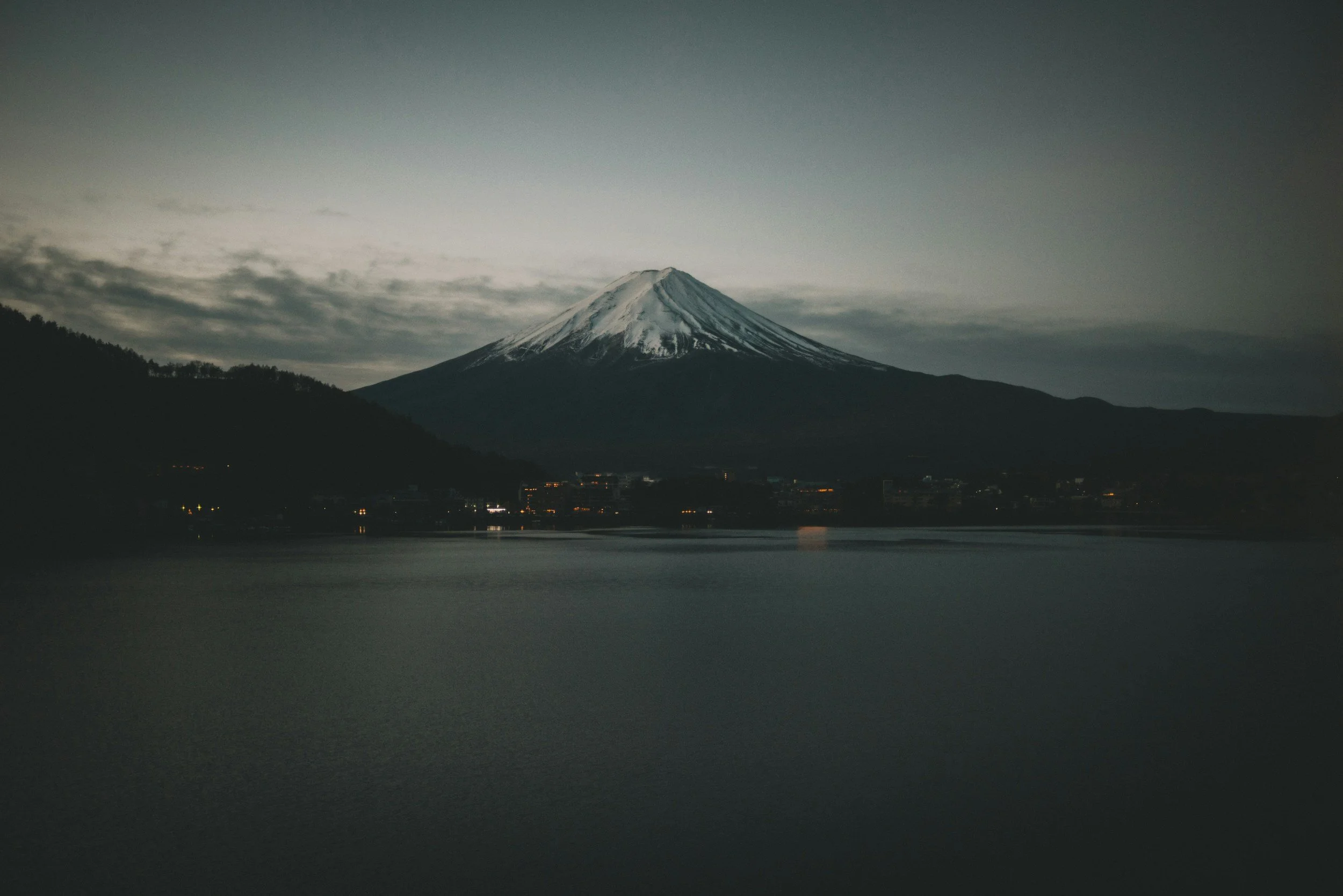Snow-capped mountain, likely Mount Fuji, viewed across a body of water during dusk or early evening.