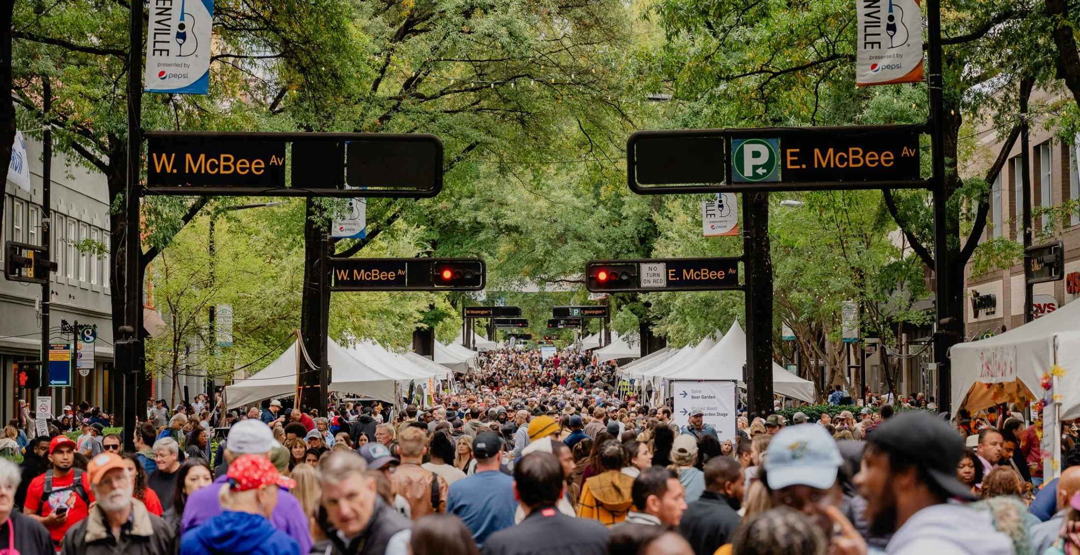Crowd of people walking along a street festival lined with white tents and trees. Signs indicate W. McBee Avenue and E. McBee Avenue, with some traffic lights and banners overhead.