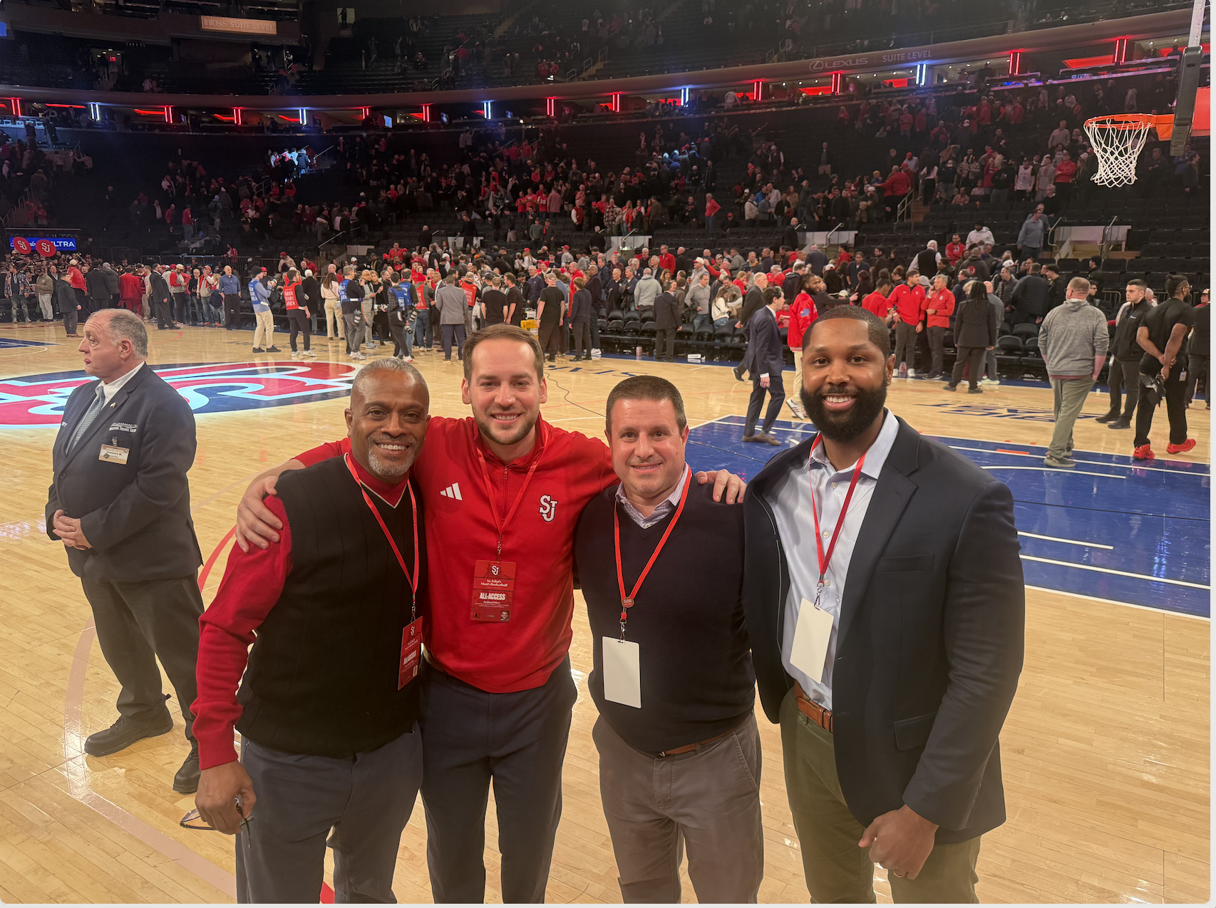 Four men smiling and posing for a photo on a basketball court with a crowd in the background. They are wearing badges and some are in jackets or sweaters, with some wearing red clothing. The background features a basketball hoop, a crowd of spectators, and people standing or walking around on the court.