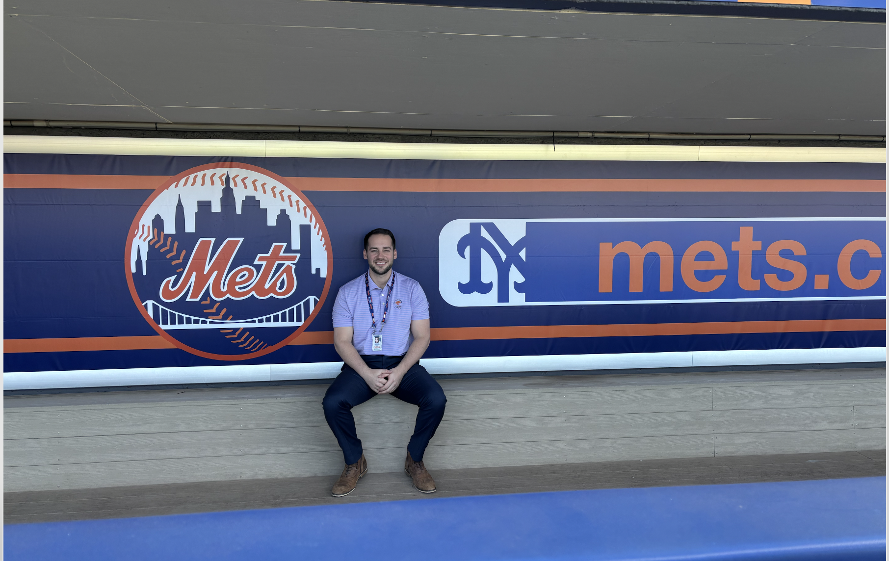 A man sitting in front of a New York Mets promotional banner at a stadium or event. The banner features the Mets logo and website address.