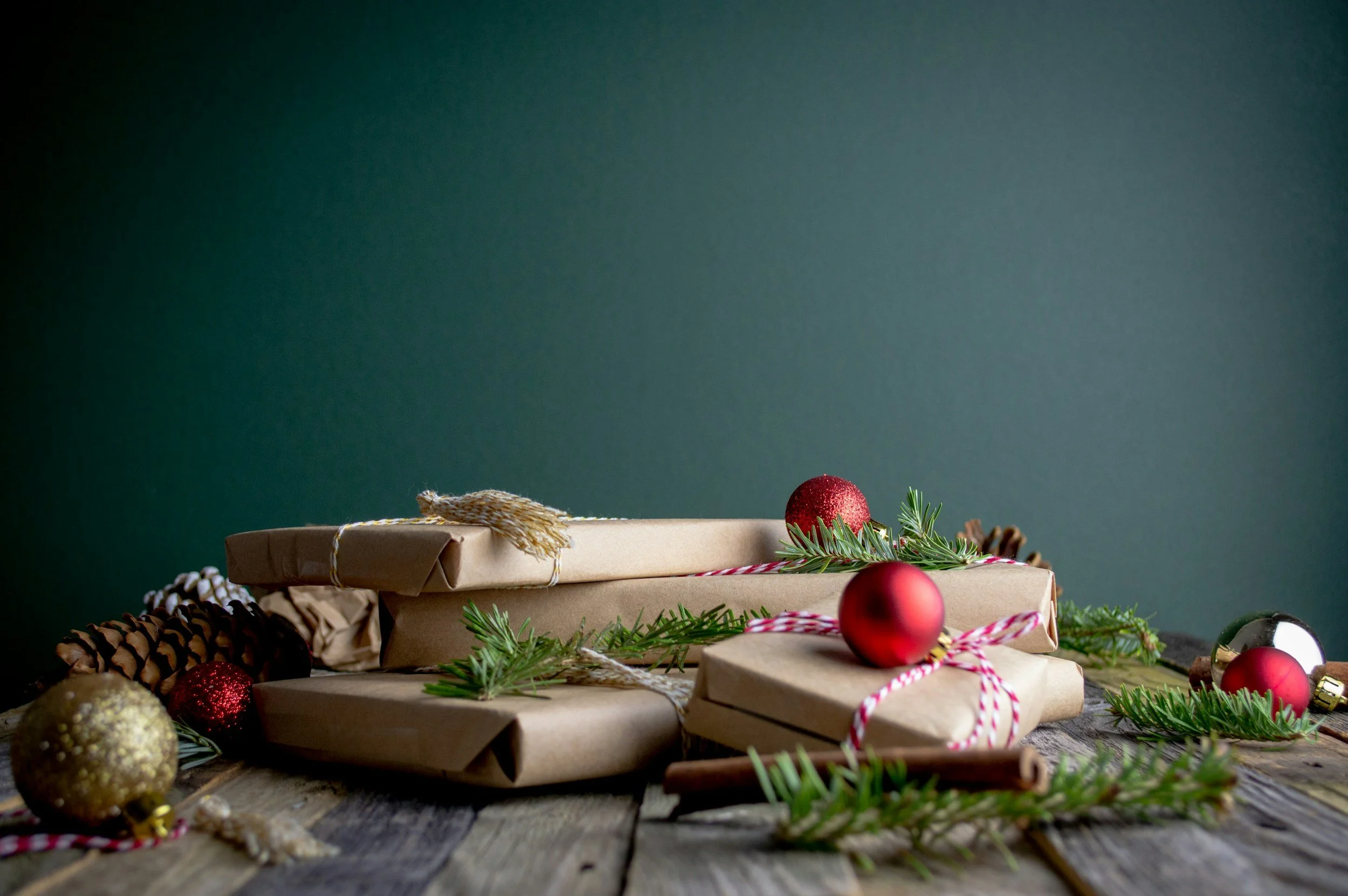 Christmas presents wrapped in brown paper with red and white string, decorated with pine branches, red ornaments, and cinnamon sticks on a rustic wooden table against a green background.