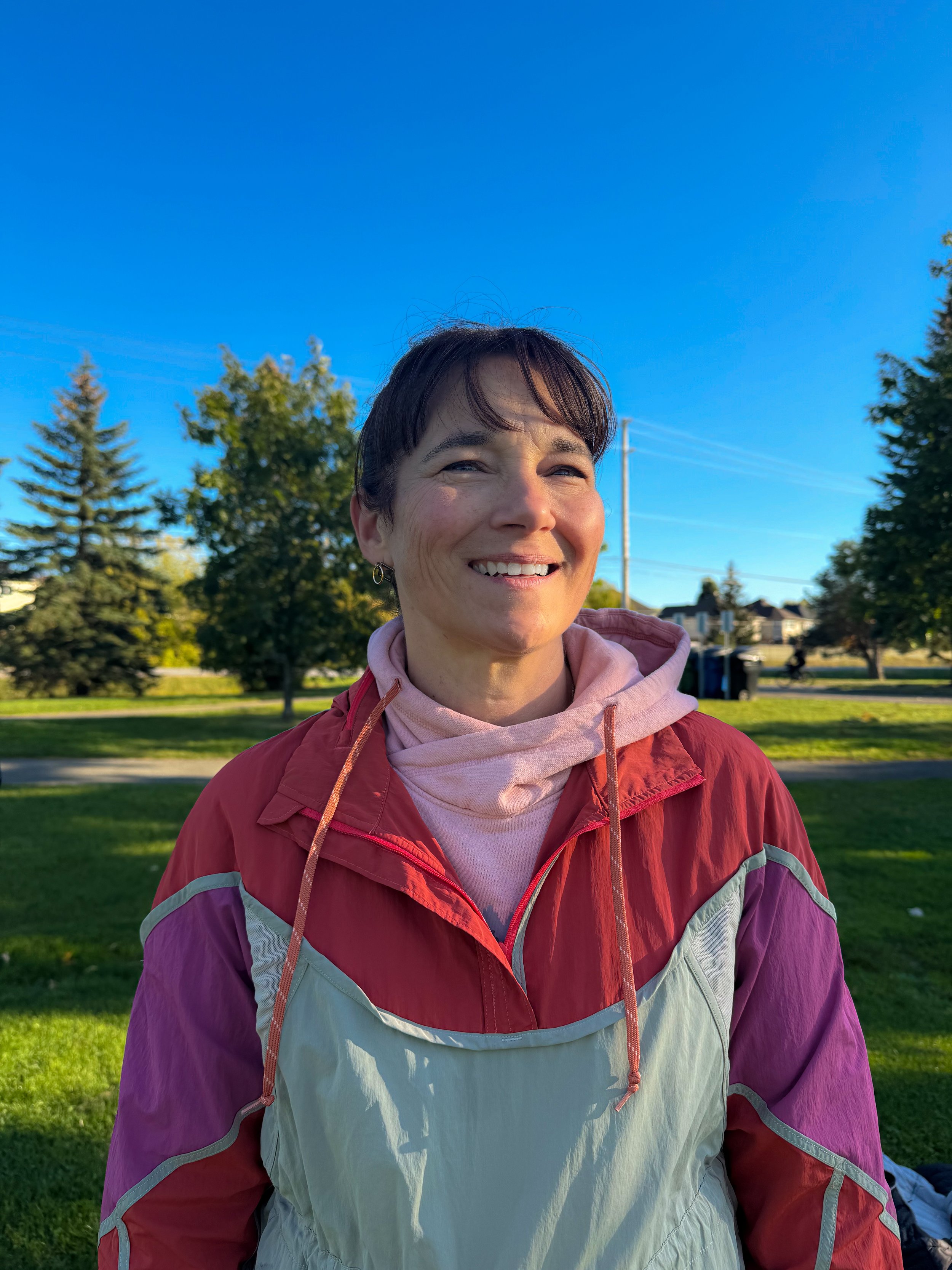 Smiling woman with short brown hair, wearing a pink and purple jacket, standing outside in a park with green trees and houses in the background under a clear blue sky.