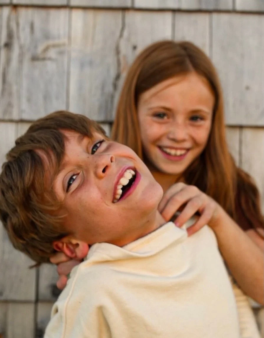 Two children, a boy and a girl, smiling and laughing together outside against a wooden background.