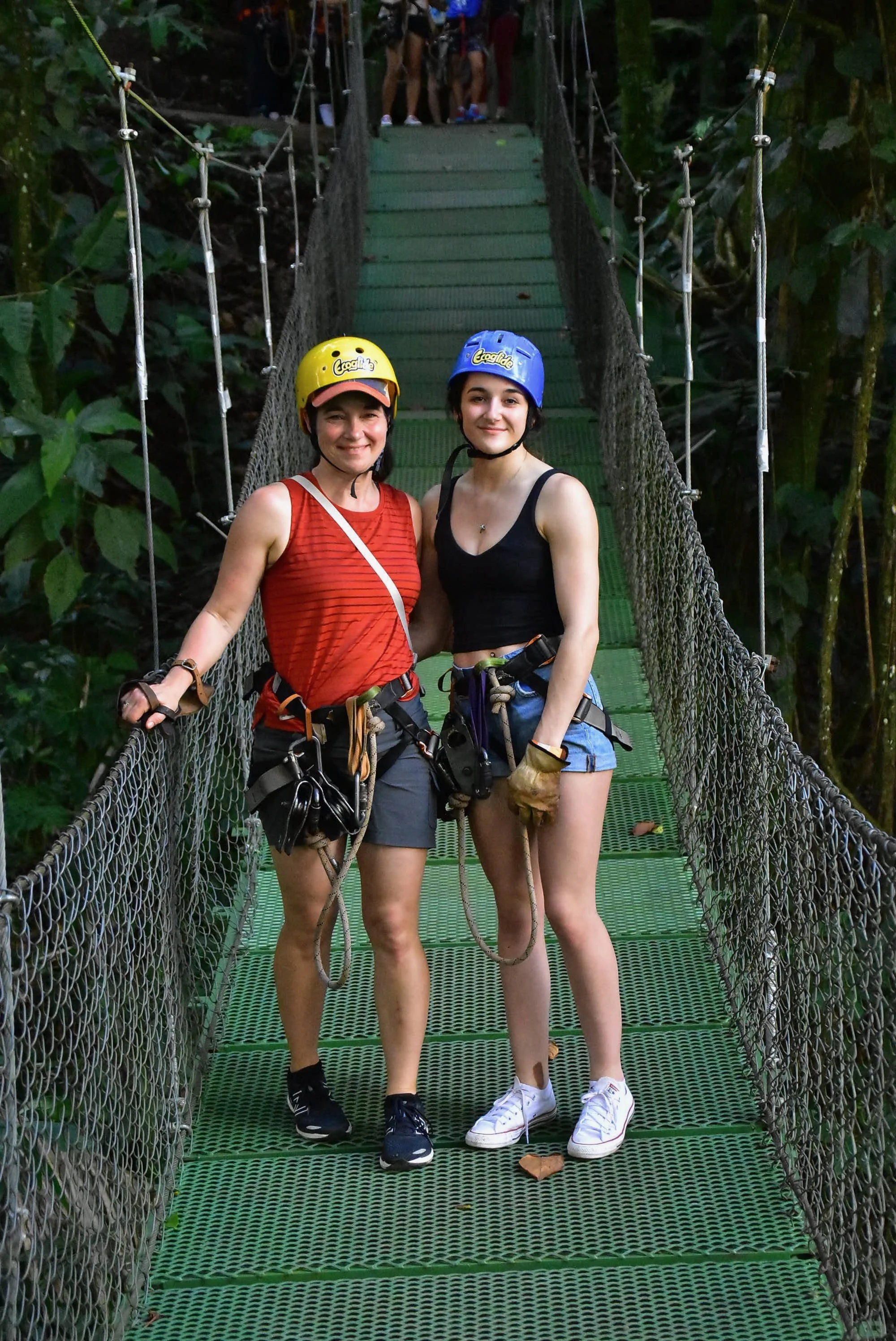 Two women wearing helmets and harnesses standing on a suspension bridge in a lush, green jungle.
