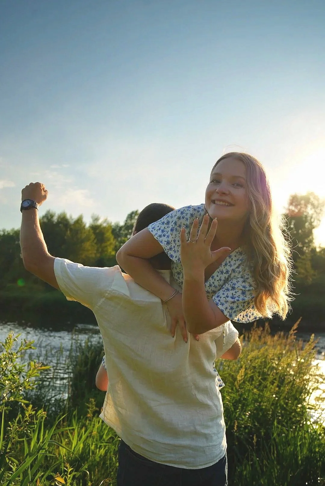 A woman is being carried on a man's shoulders outside by a body of water during sunset, smiling and looking at the camera.