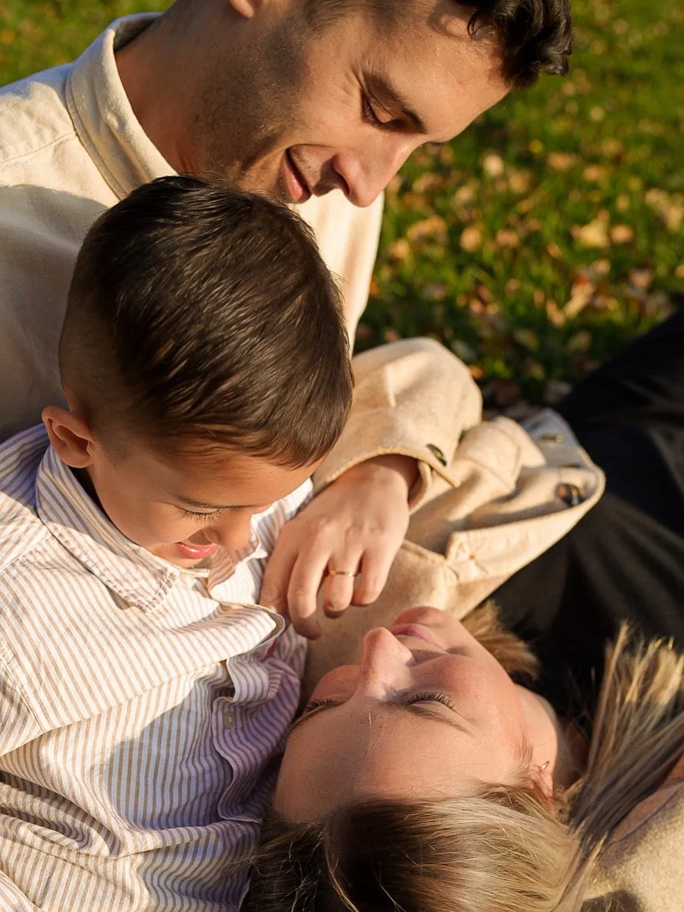 A family of three lying on the grass during sunset, smiling, with the father and mother looking at their young son, capturing a joyful moment.