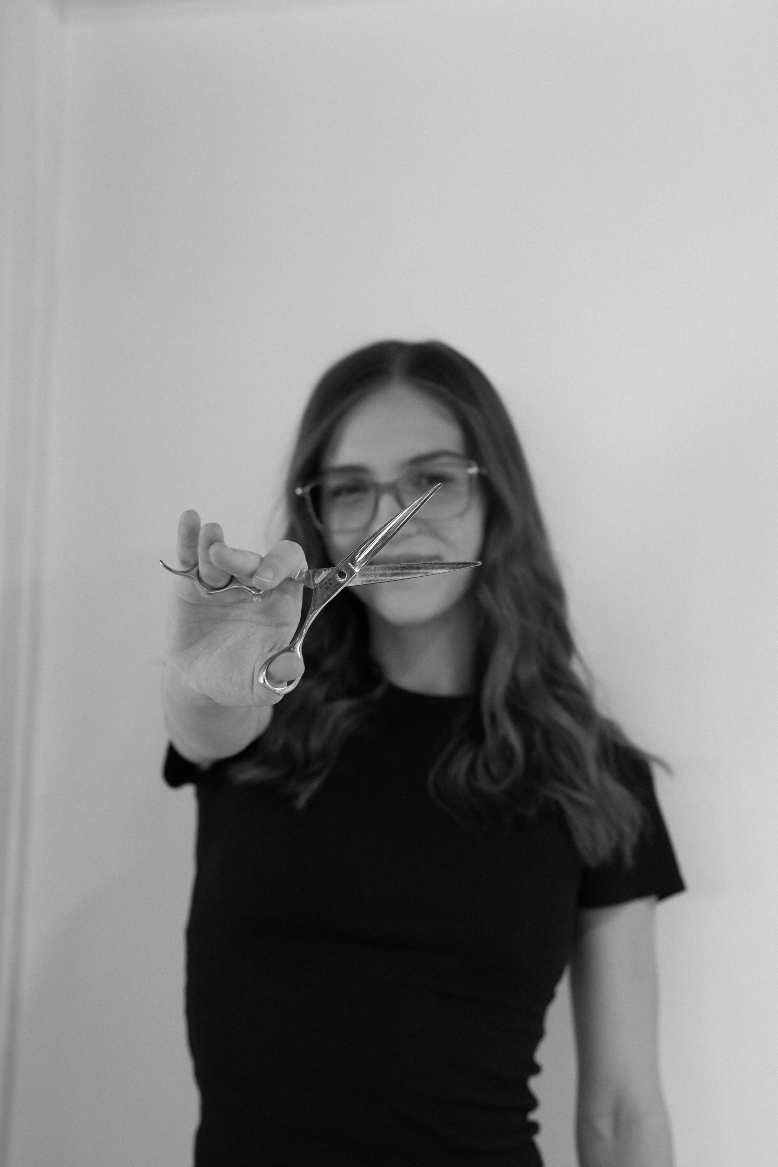 Woman with long hair and glasses holding scissors towards the camera in front of a plain wall.