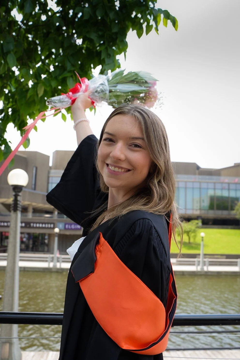 A young woman in a graduation gown holding a bouquet of pink flowers, smiling outdoors near a body of water with a modern building in the background.