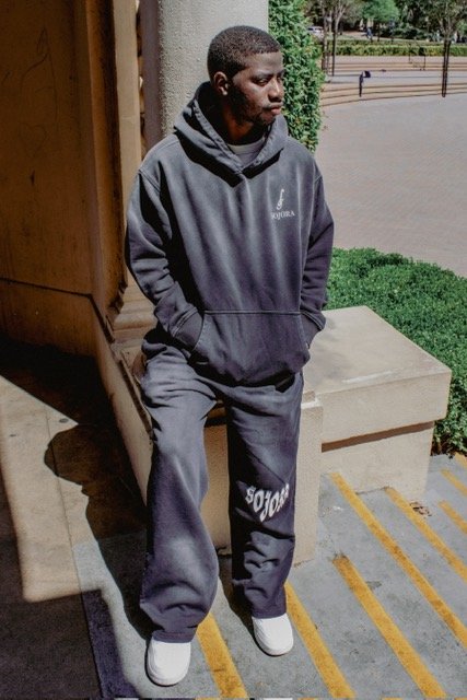 A young man in a black hoodie and sweatpants standing outdoors by a building, with hands in hoodie pockets and looking down.