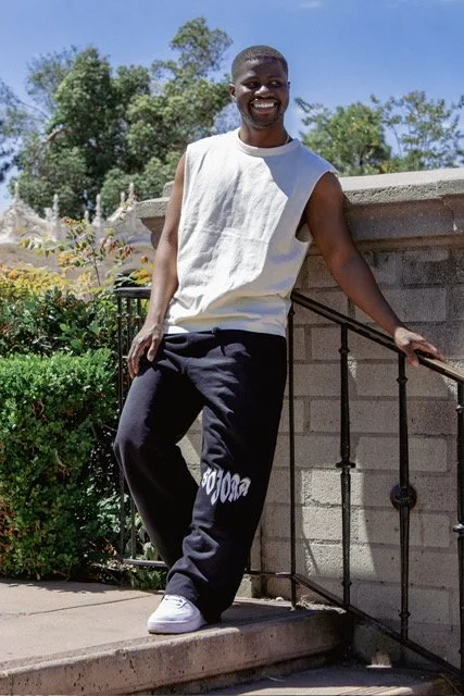 A smiling young man standing outdoors on steps made of concrete, wearing a sleeveless white shirt, black sweatpants, and white sneakers, with greenery and trees in the background.