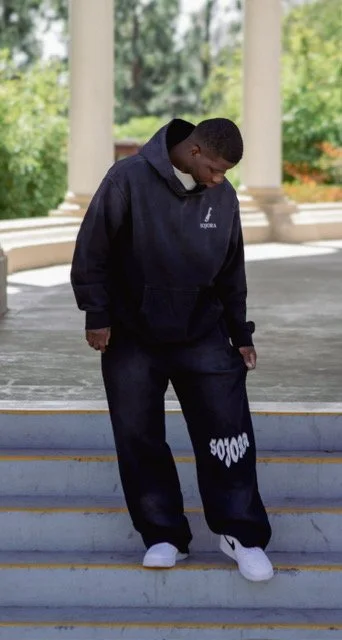 A young man standing on a staircase outdoors, dressed in a black hoodie and sweatpants, with white sneakers, and looking down.