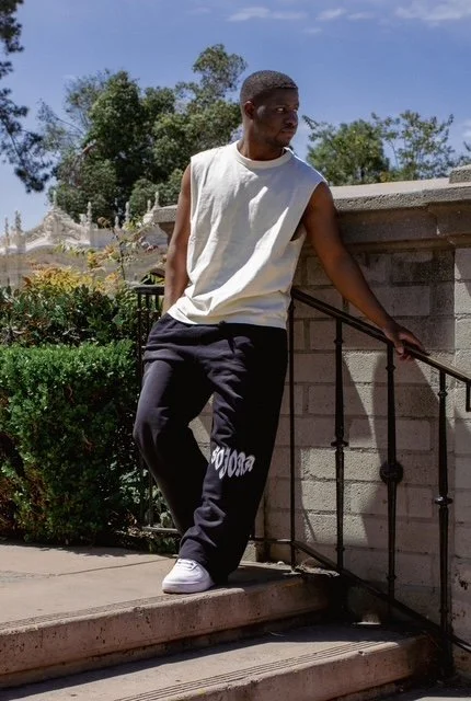 Young man standing on outdoor steps, leaning on a black metal railing, wearing a sleeveless white shirt, black sweatpants, and white sneakers, with trees and a blue sky in the background.