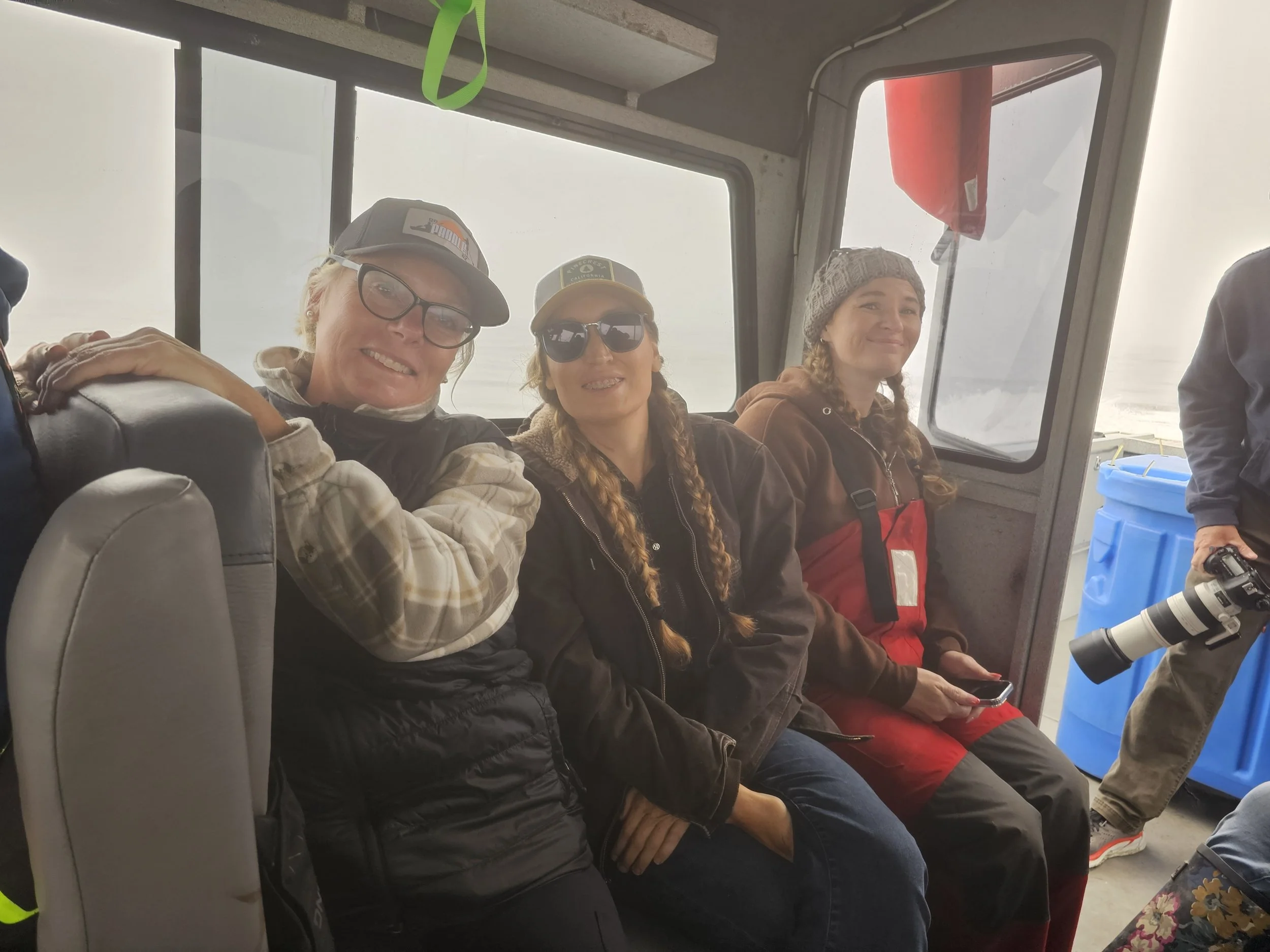 Three women sitting inside a boat, smiling, with fishing gear, and a person standing with a camera and a large blue container in the background.