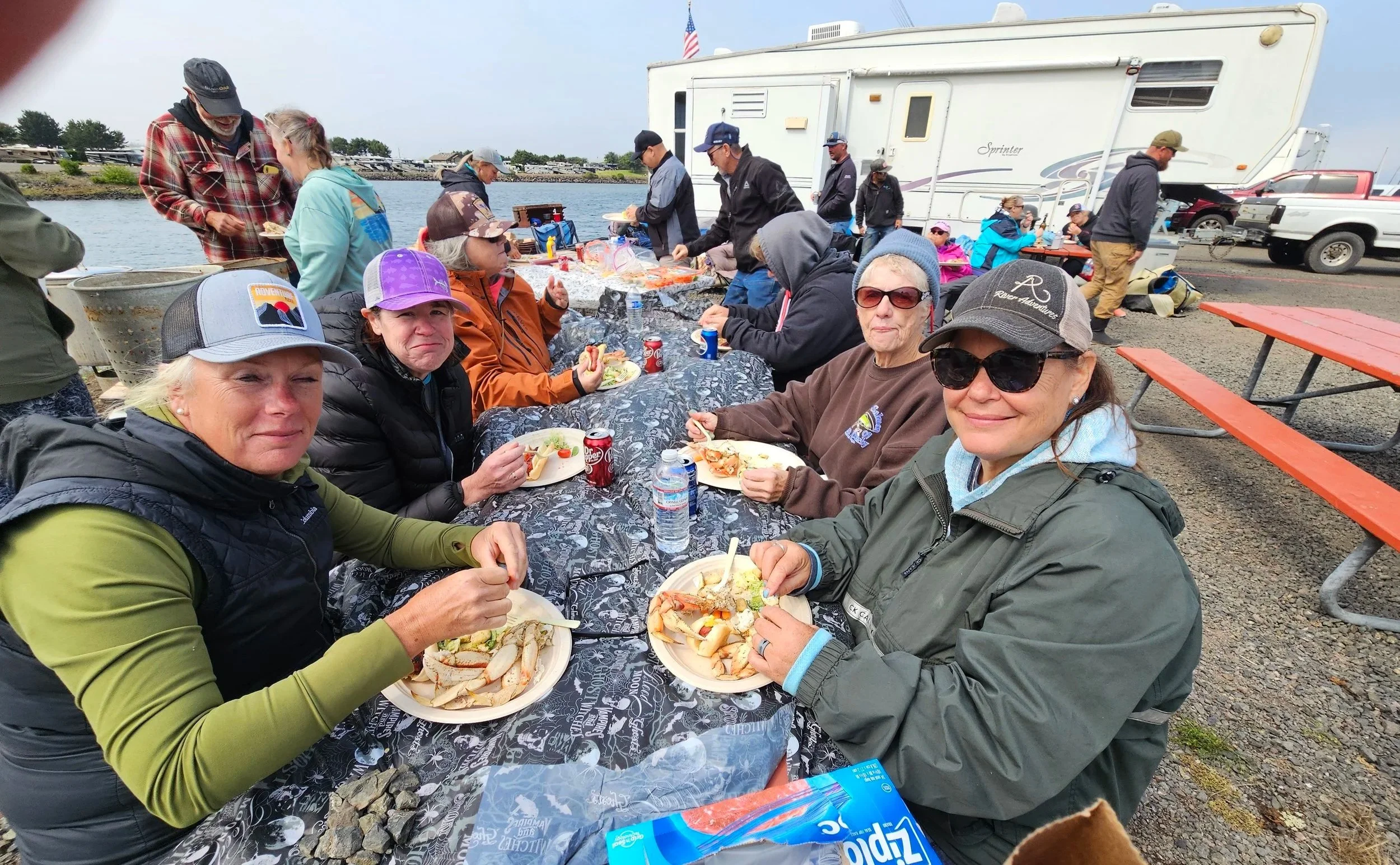 People sitting at a long outdoor table enjoying a meal, with some eating seafood, near parked RVs and vehicles by a body of water on cloudy day.