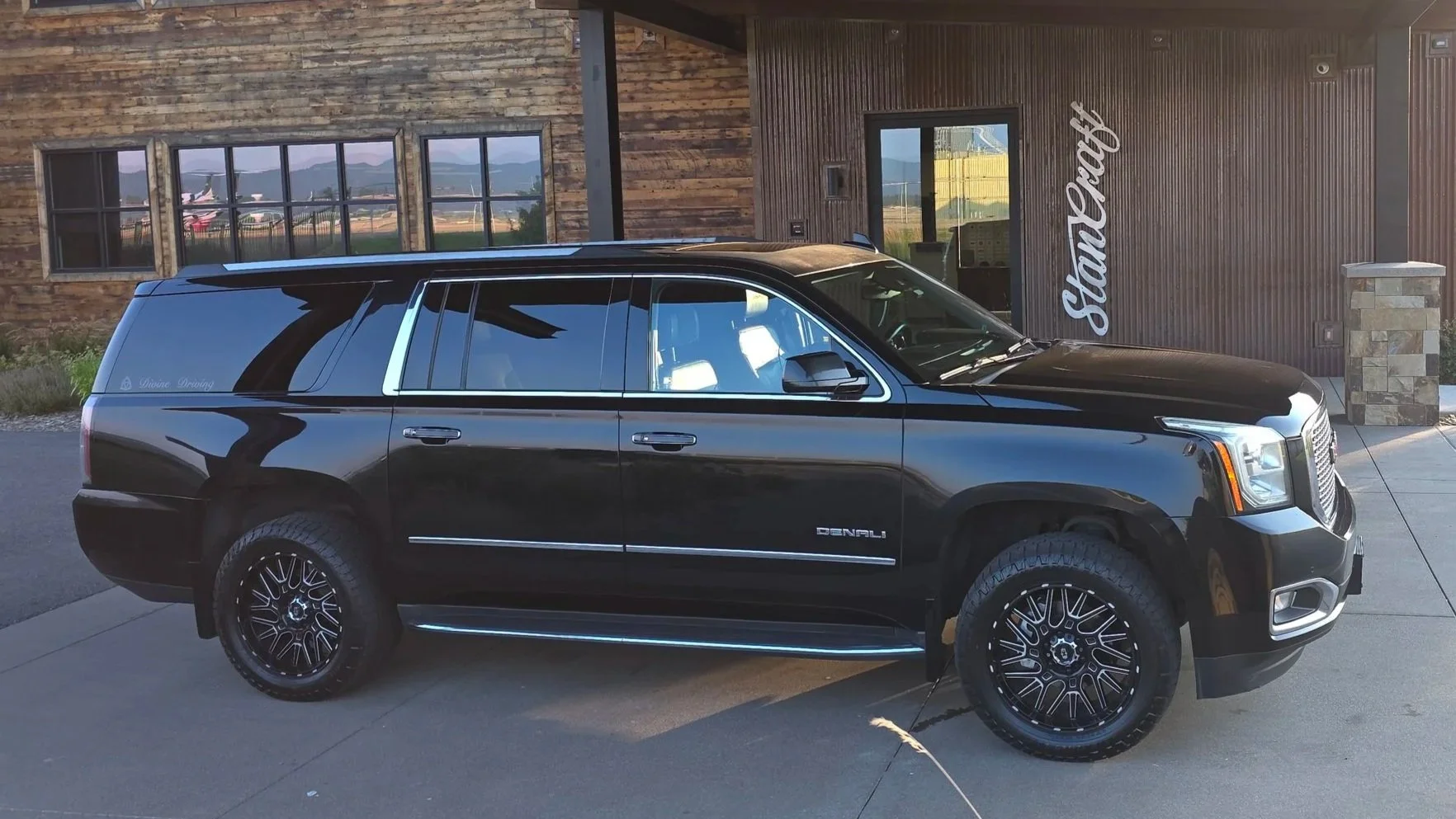 A black GMC Yukon XL Denali SUV parked in front of a private airport building with wooden siding and a sign that reads 'Stancraft'. The building has large windows and a stone pillar at the entrance. Mountain scenery is reflecting in the windows.