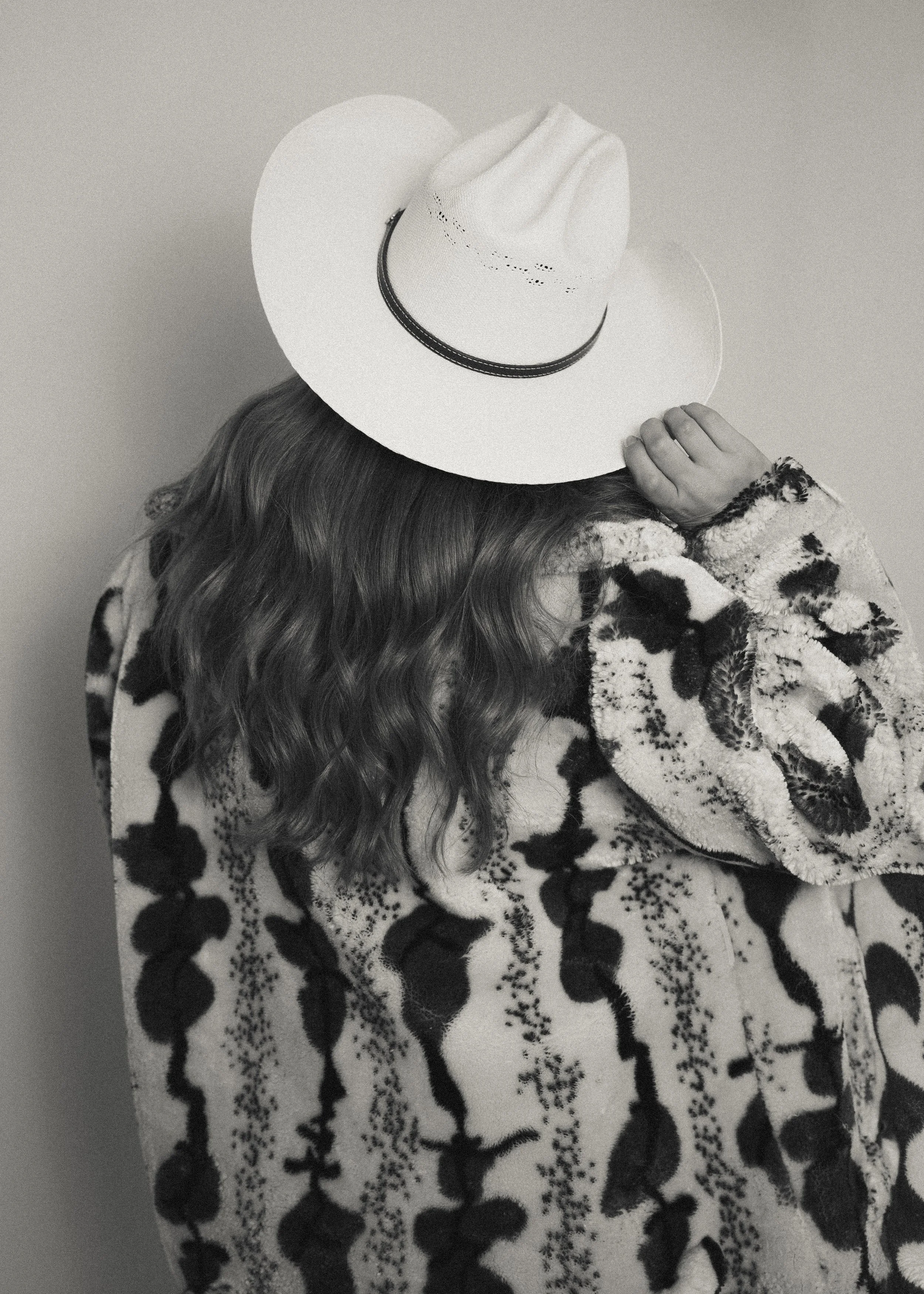 A person with long, curly hair is wearing a wide-brimmed white hat with a black band. They are wearing a patterned, fuzzy, long-sleeved top with animal print. The person's face is not visible, and they are touching the side of the hat with one hand.