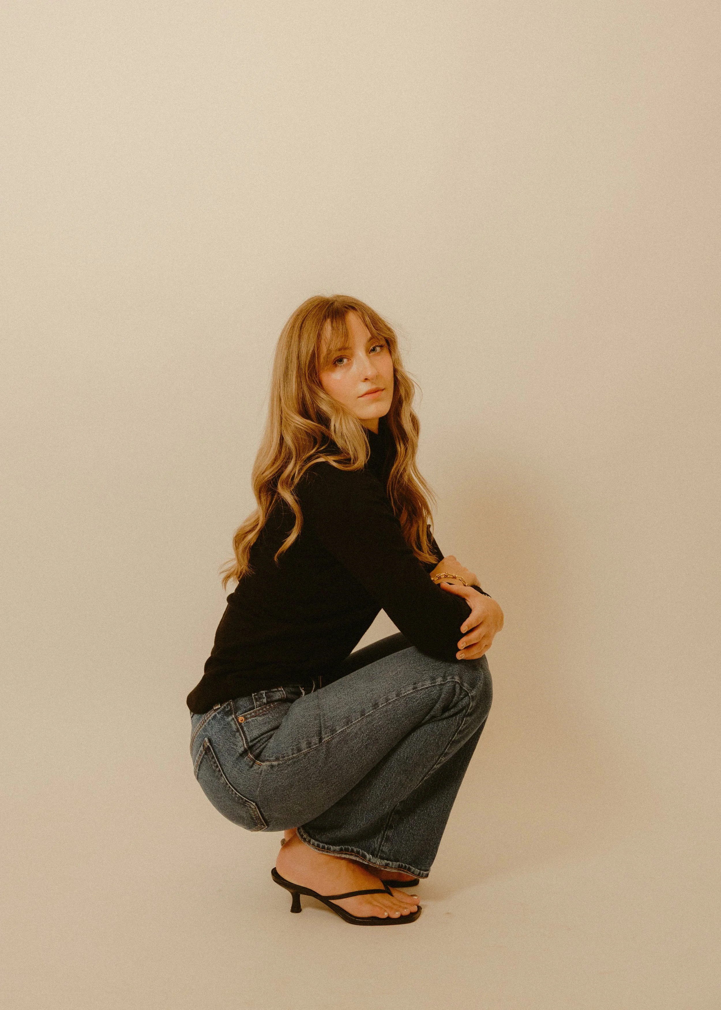 A young woman with long, wavy blonde hair and light skin posing against a plain beige background. She is wearing a black long-sleeve top, denim jeans, and black high-heeled sandals. She has a confident expression, with one knee raised and her arms crossed over her thighs.