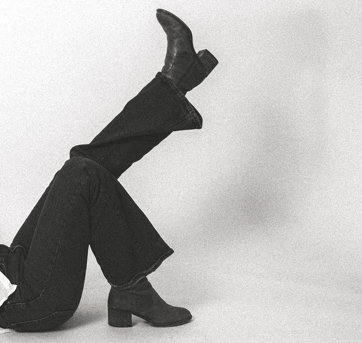 Black and white photo of a person wearing high-heeled boots and jeans, balancing on their hands with their legs extended upward.