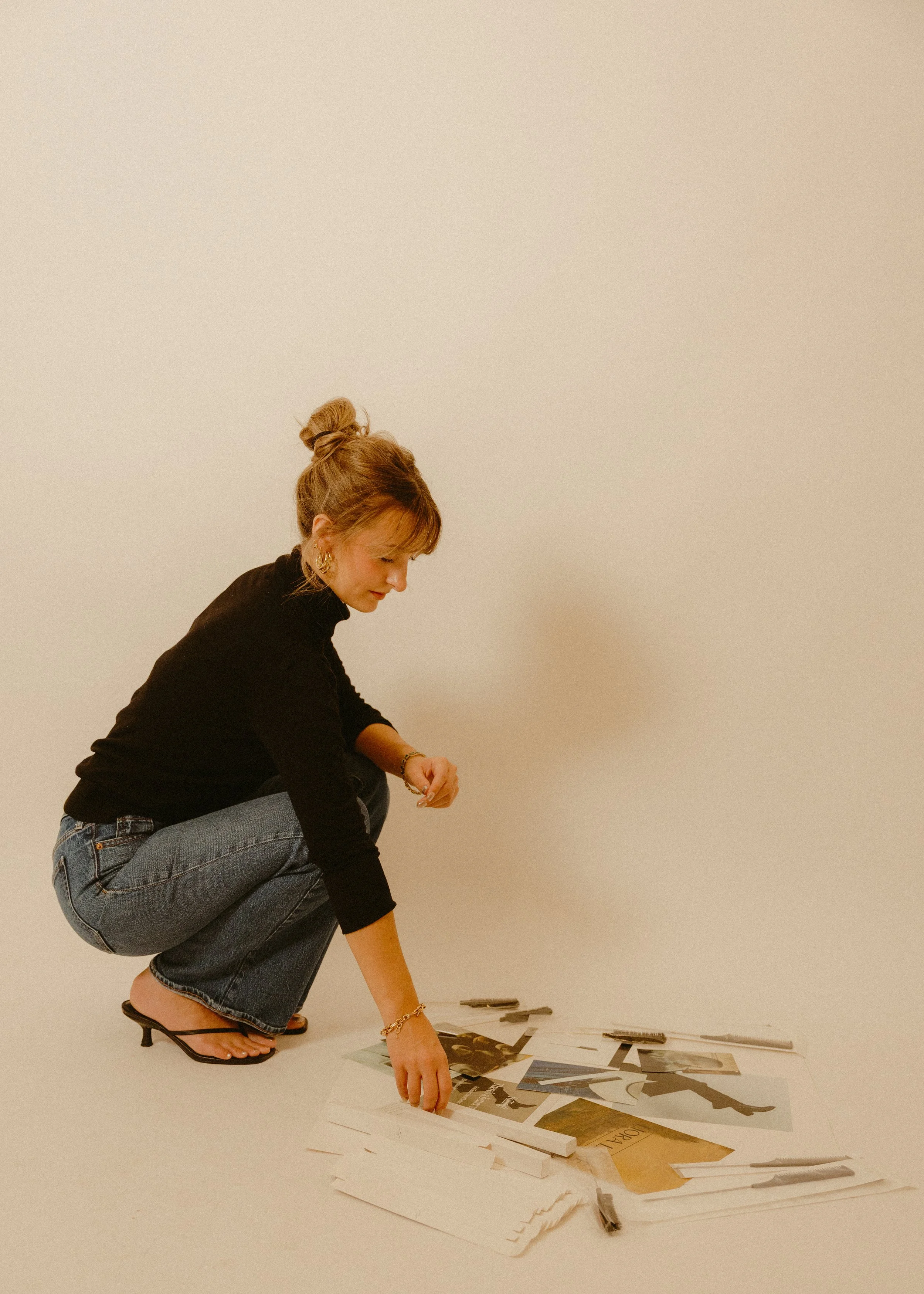 A woman crouched down, wearing a black sweater, jeans, and high-heeled sandals, arranging photographs or artwork on the floor against a plain, light-colored wall.