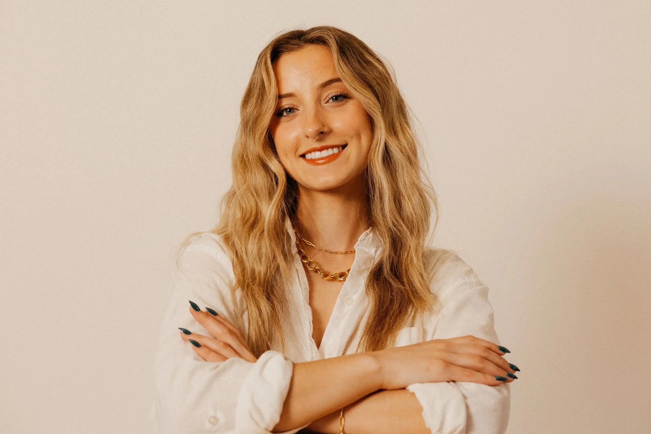 A young woman with wavy blonde hair and fair skin, smiling with her arms crossed, wearing a white shirt, gold chain necklace, and a nose ring against a plain beige background.
