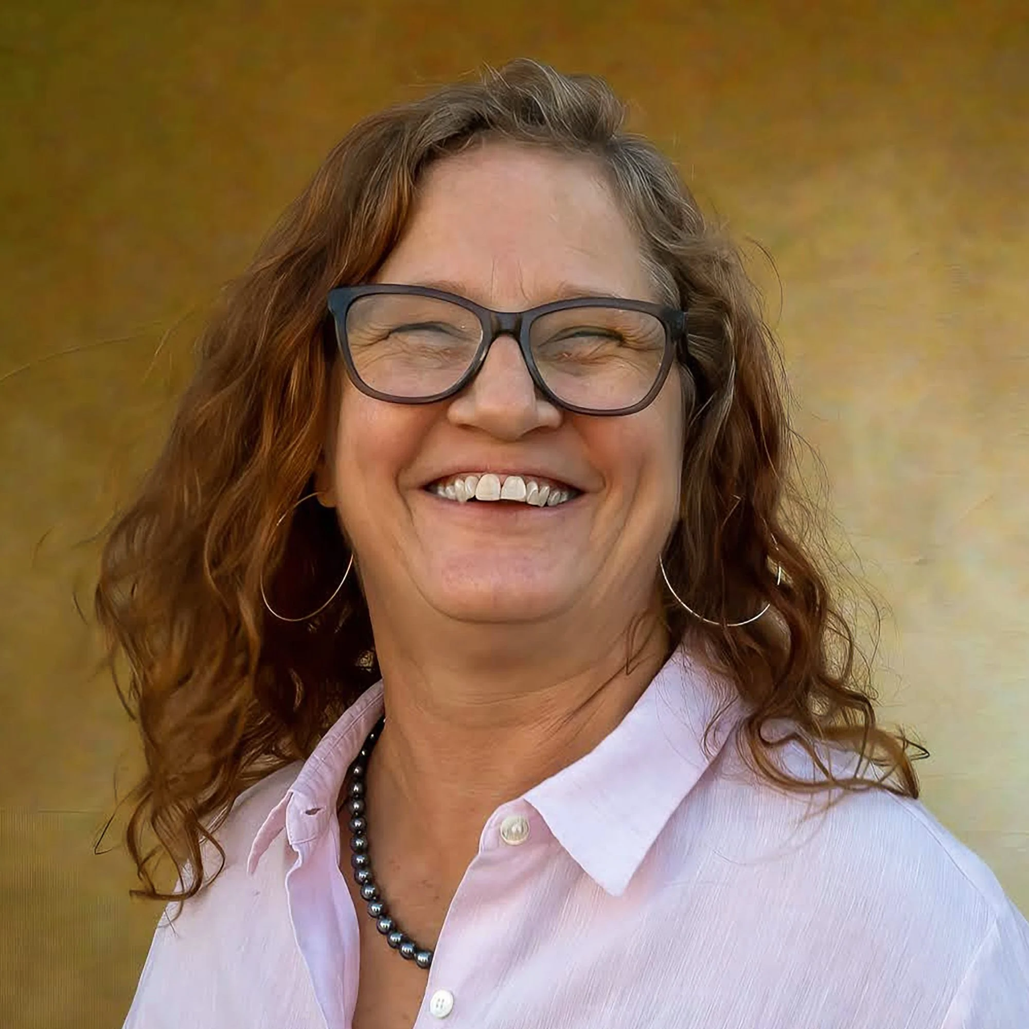 A woman with curly red hair wearing glasses, a pink button-up shirt, a black necklace, and hoop earrings, smiling.