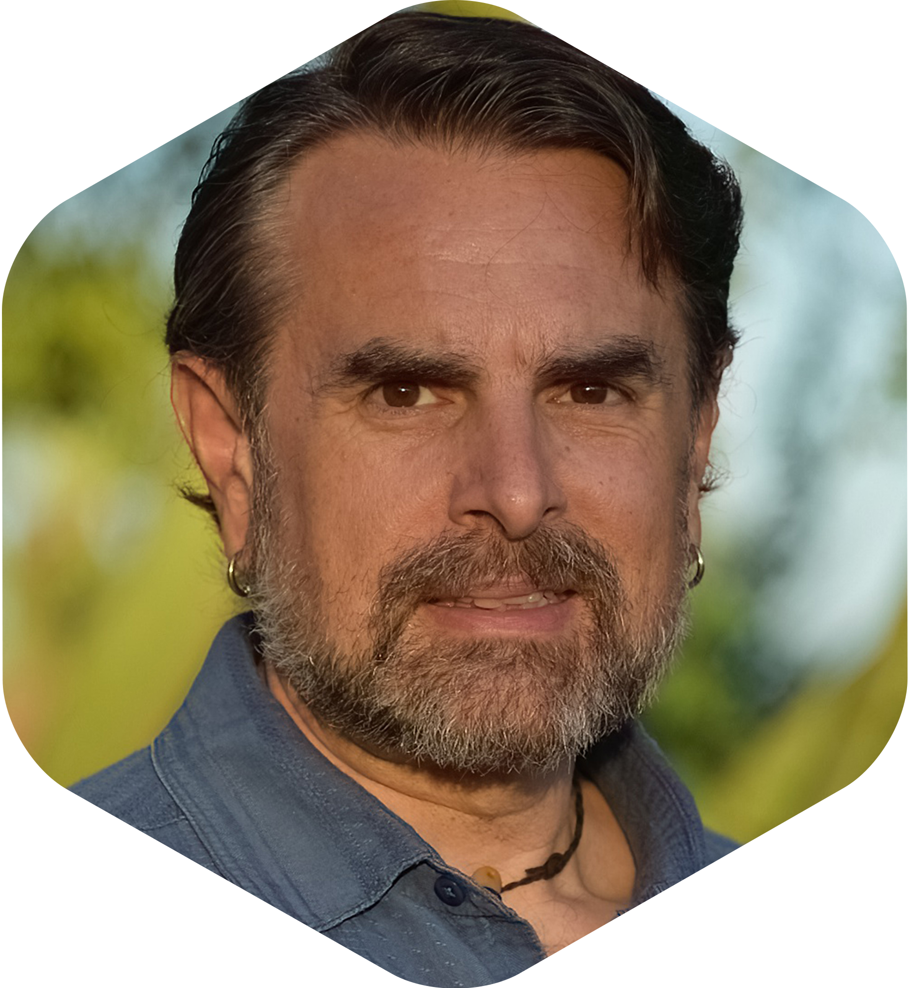 Sean Ellis has dark hair, brown eyes, and a beard, wearing a dark blue shirt and earrings, outdoors with blurred green and yellow background.