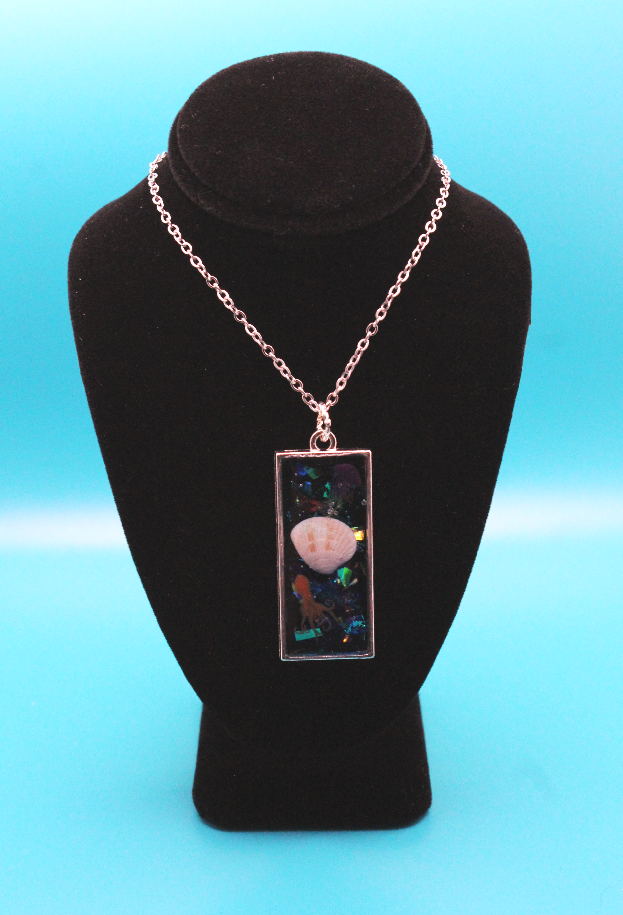 Silver rectangle pendant filled with glittery blue resin - set inside are an orange resin octopus, a real seashell, and a resin yellow and purple jellyfish. It is strung on a silver chain.
