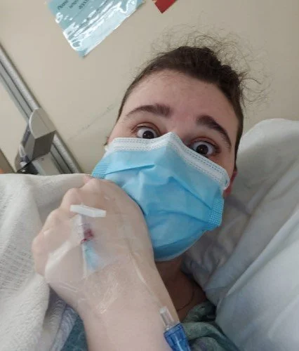A person with brown, frizzy hair shows off the IV on the back of their hand, wearing a medical mask in an ER