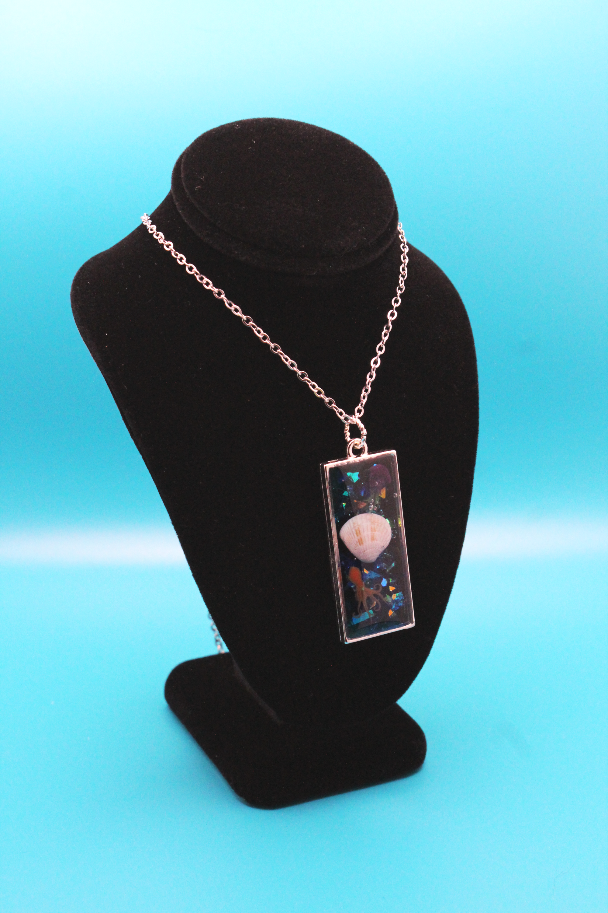 Silver rectangle pendant filled with glittery blue resin - set inside are an orange resin octopus, a real seashell, and a resin yellow and purple jellyfish. It is strung on a silver chain.