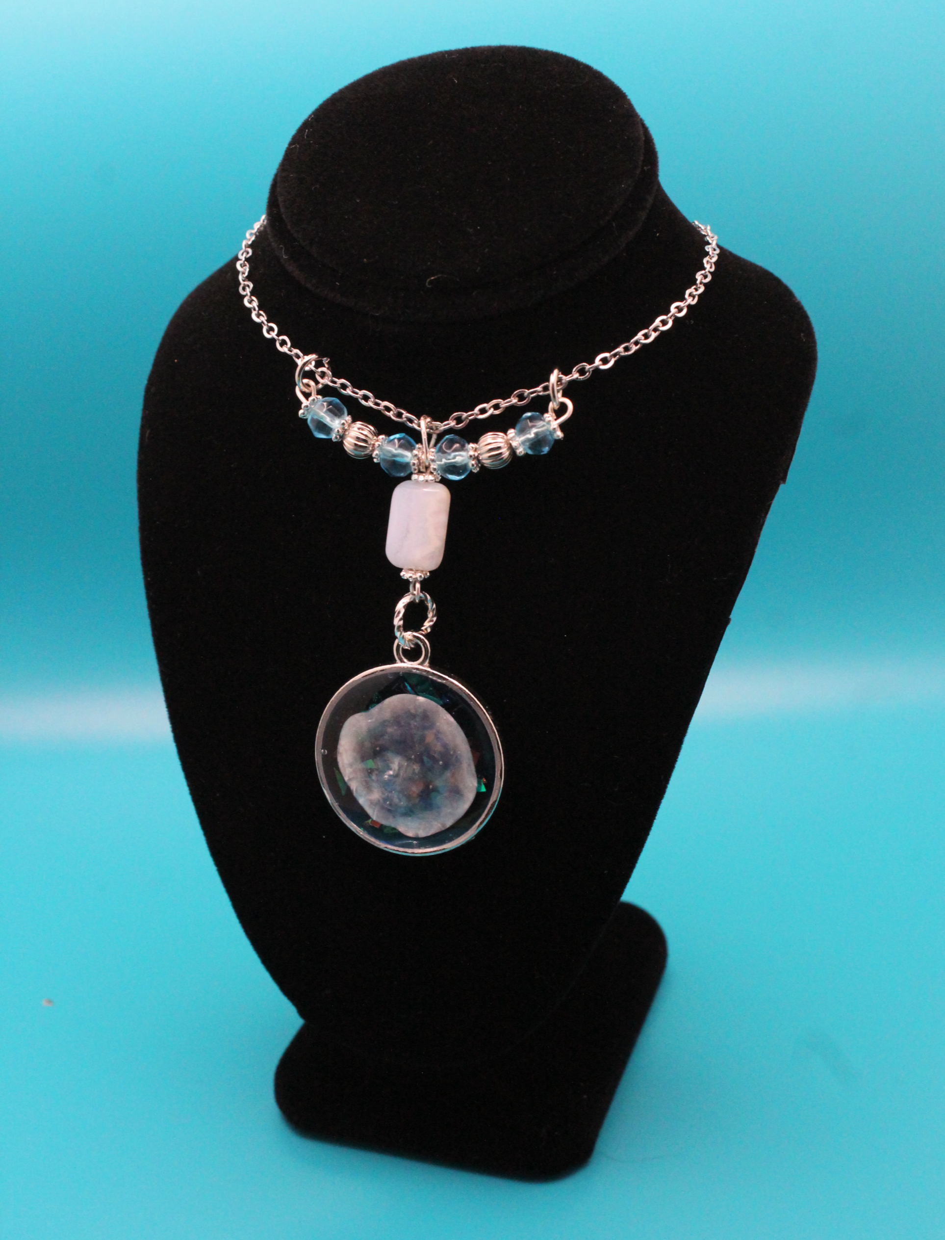 silver pendant with a seashell set in blue and glitter resin, hanging from a rectangular stone that's strung with blue and silver beads on a silver chain