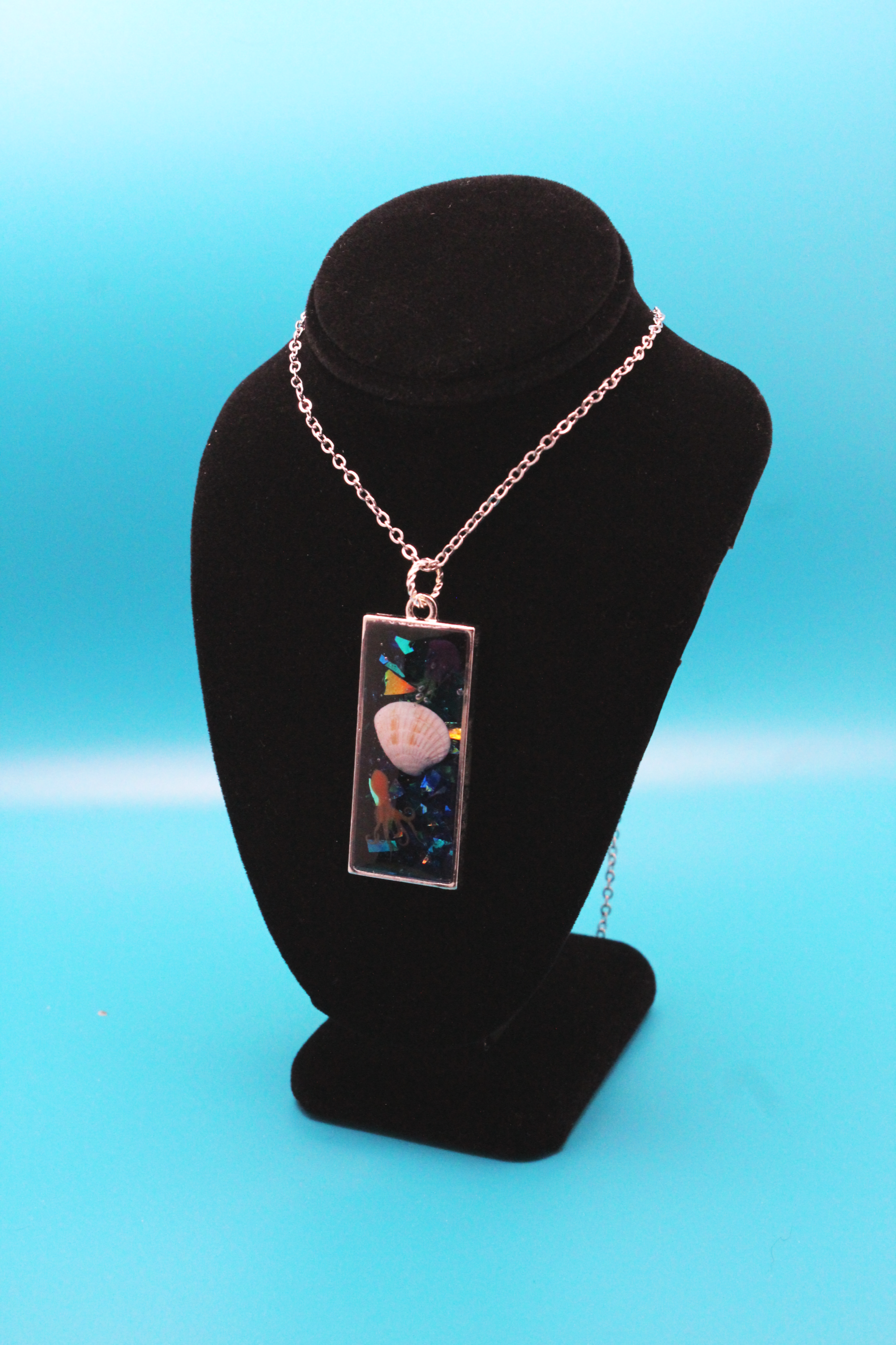 Silver rectangle pendant filled with glittery blue resin - set inside are an orange resin octopus, a real seashell, and a resin yellow and purple jellyfish. It is strung on a silver chain.