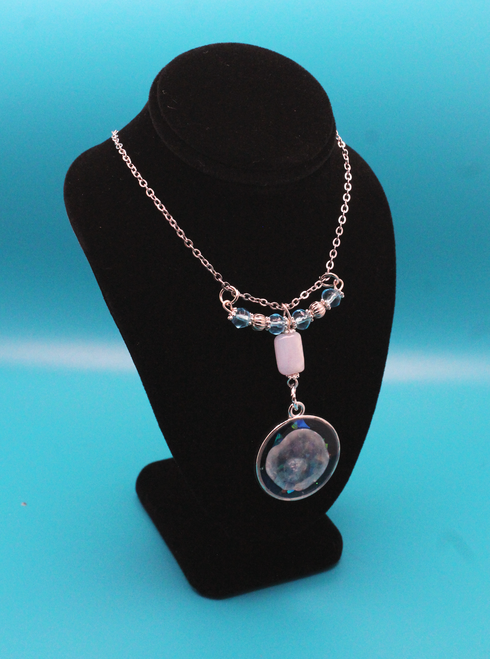 silver pendant with a seashell set in blue and glitter resin, hanging from a rectangular stone that's strung with blue and silver beads on a silver chain